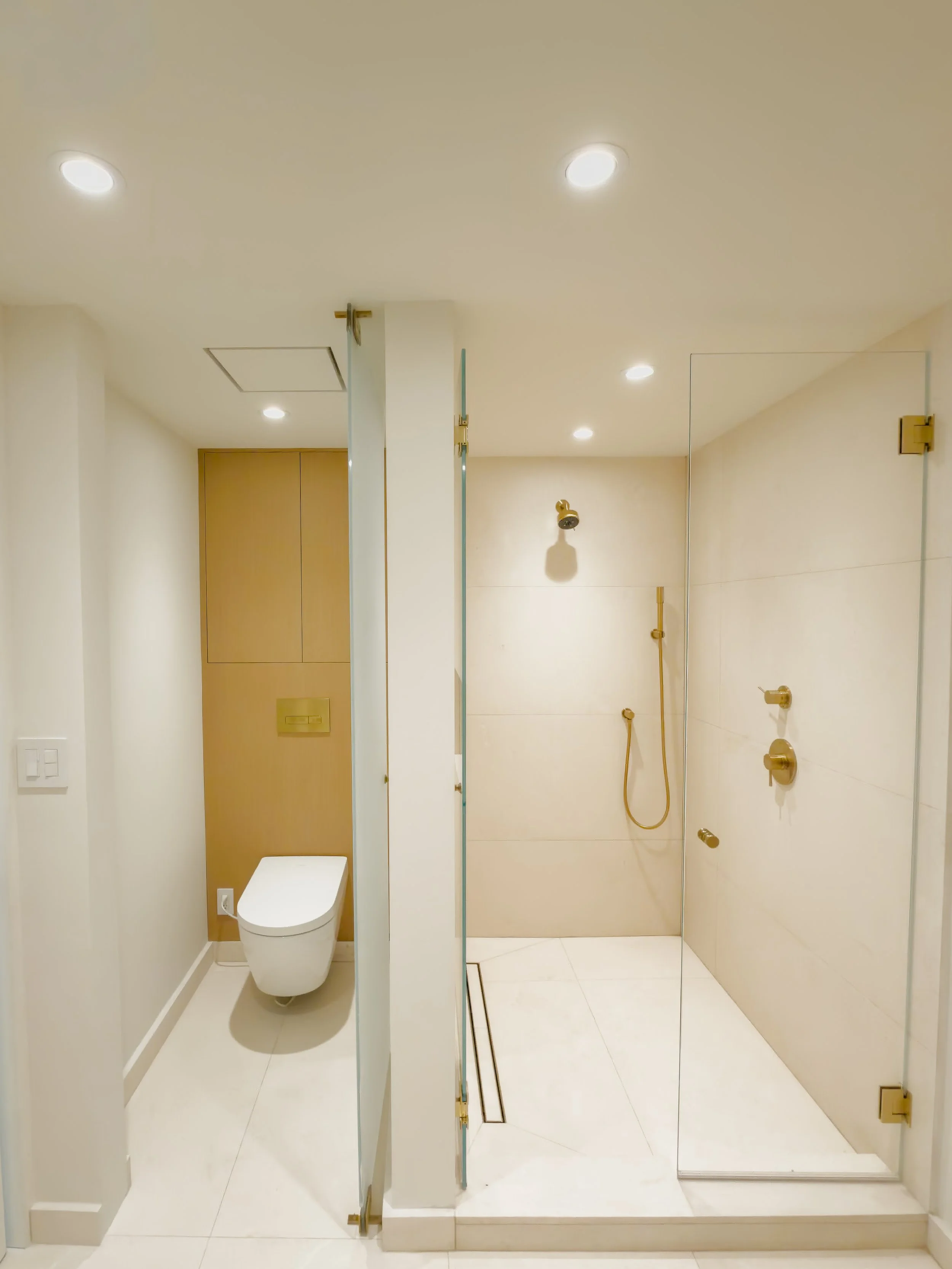 Modern bathroom with white walls and tiles, featuring a toilet, a glass shower enclosure with gold fixtures, a wall-mounted showerhead, and recessed ceiling lights.