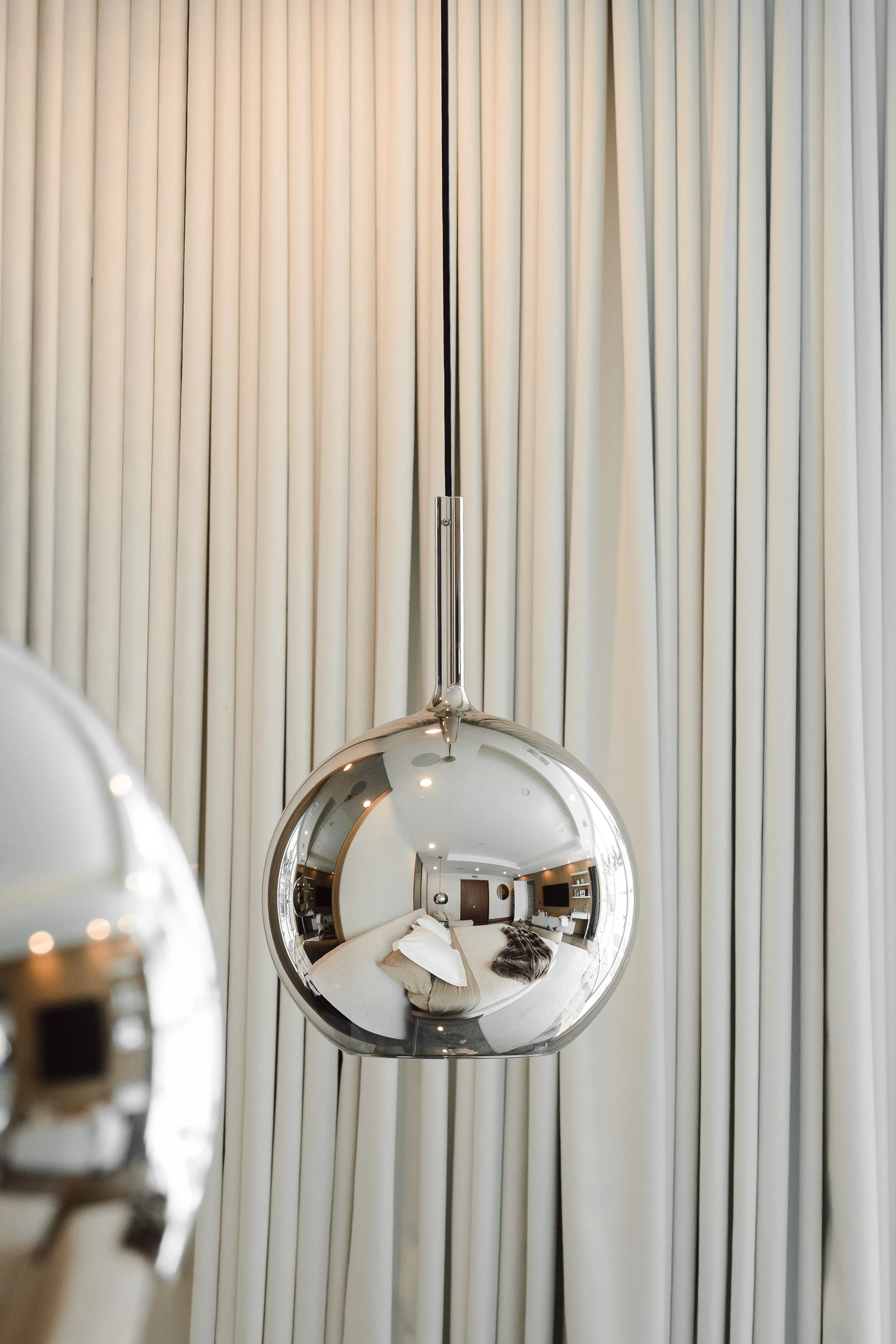Close-up of a modern chandelier with a reflective metallic sphere, reflecting a hotel room with light-colored walls, bedding, and a television.
