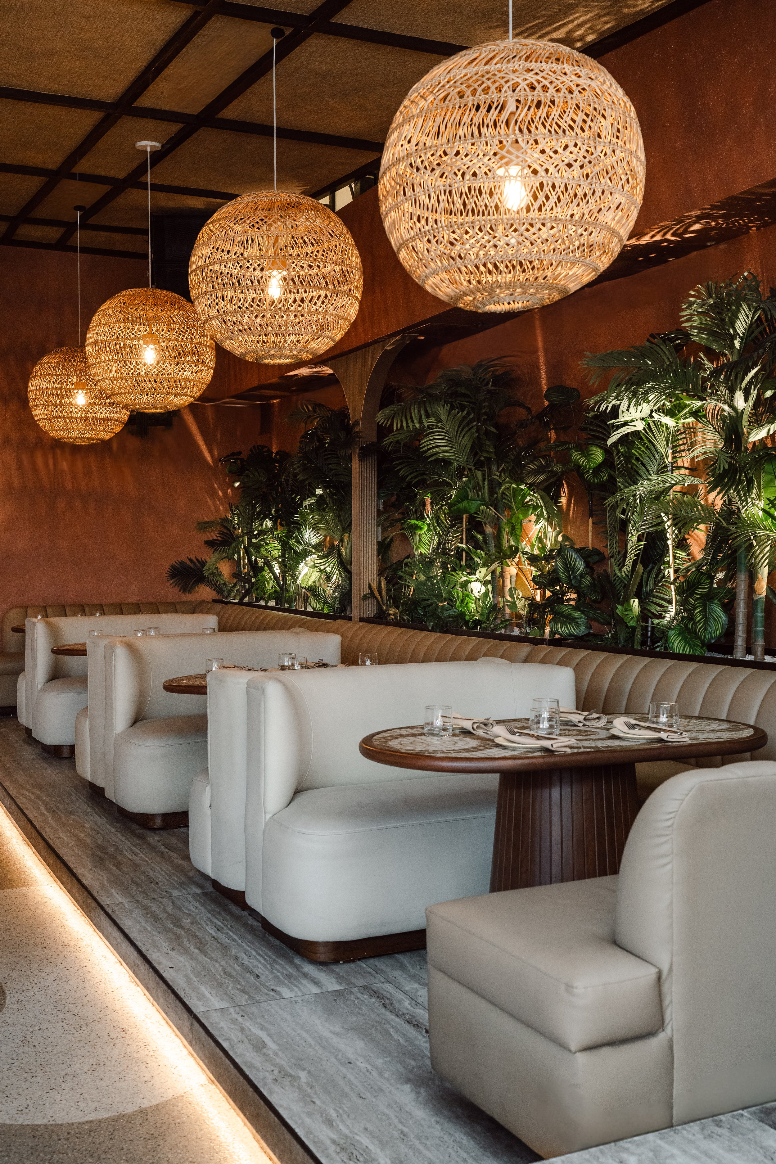 A cozy restaurant interior featuring white upholstered booths, wooden tables set with glasses and napkins, hanging wicker ball pendant lights, and lush green plants on the wall.
