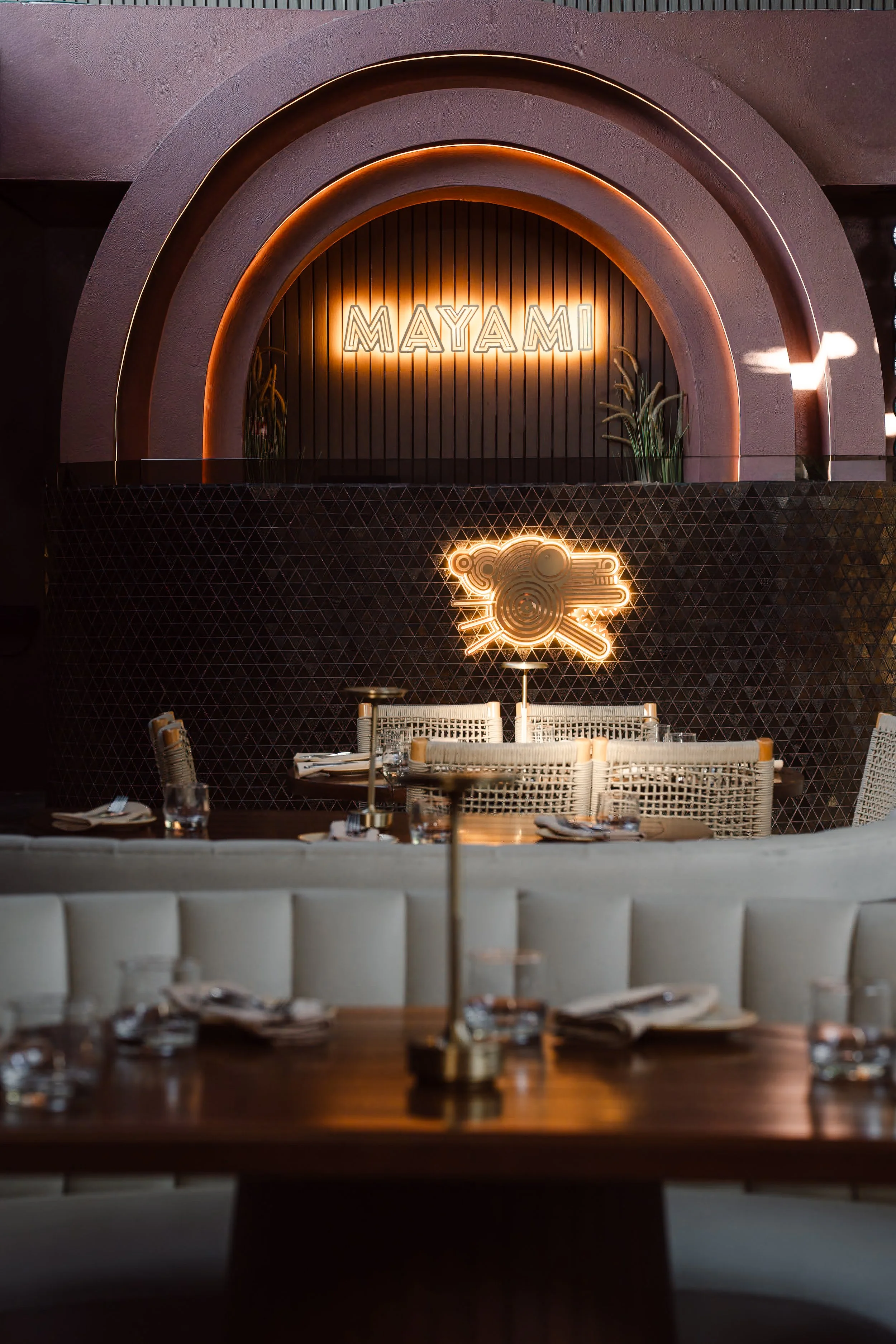 Interior view of a restaurant with a neon sign reading 'MAYAMI' on a dark, ribbed wall. There are green plants and a patterned sculpture or decoration beneath the sign. In the foreground, tables are set with glasses, napkins, and utensils, with chair