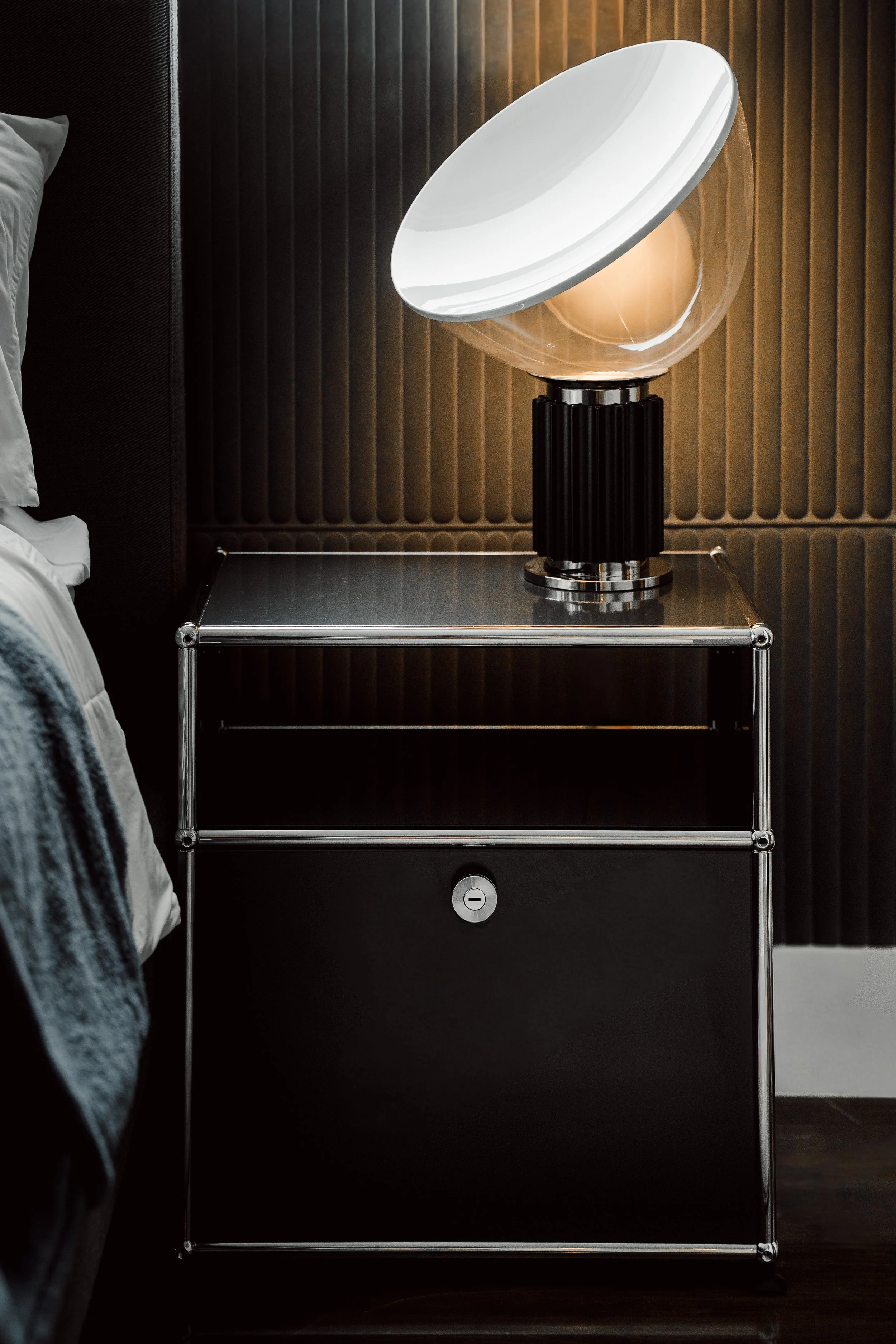 A modern bedside lamp with a spherical glass base and a white oval lampshade, placed on a black and chrome nightstand beside a bed with gray bedding.