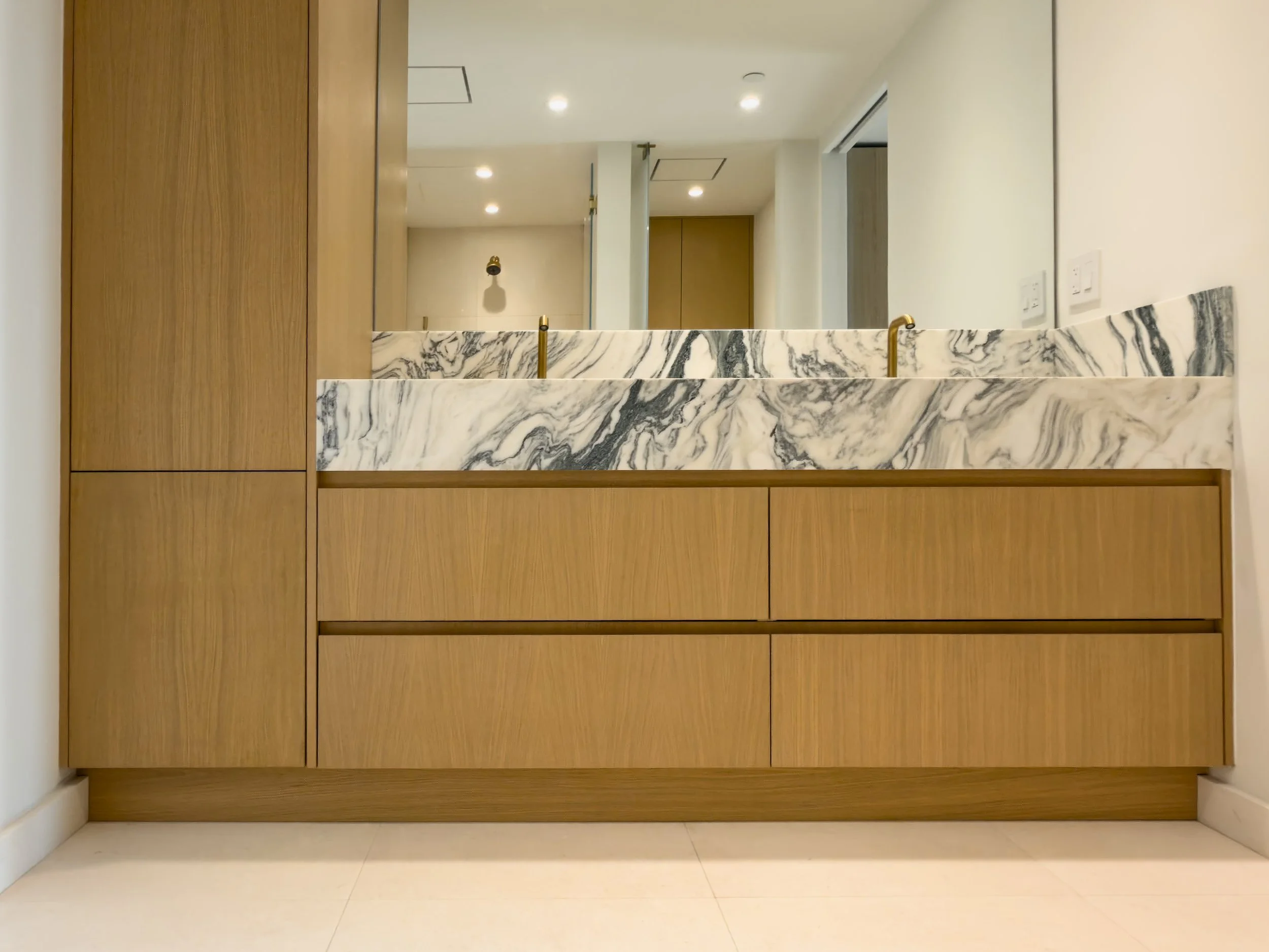 A bathroom countertop with a marble surface and two gold faucets, set against a wooden cabinet and a mirror, with a beige wall behind.