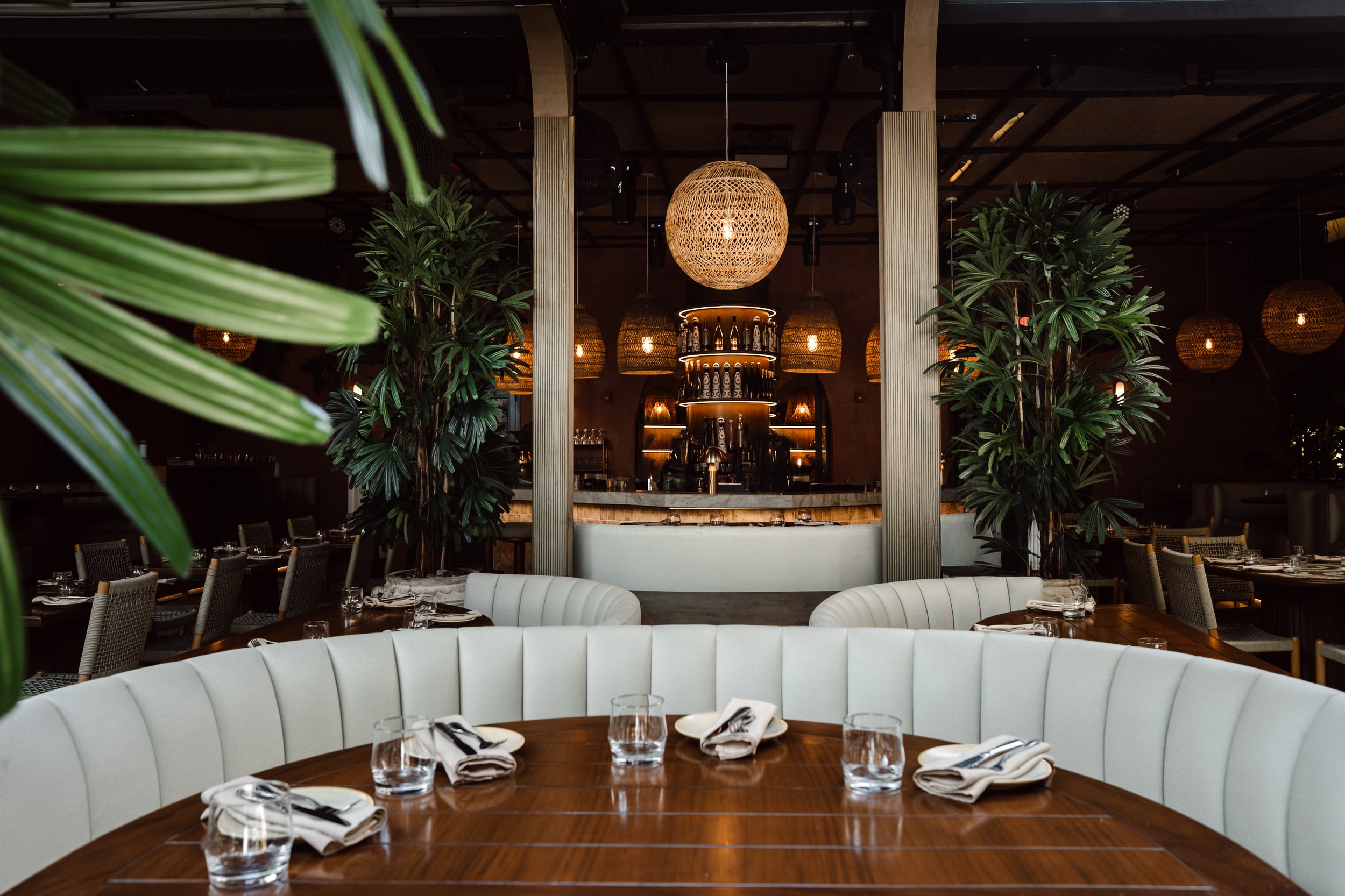 Interior of a stylish restaurant with a prominent round wooden table set with glassware and napkins in the foreground, surrounded by modern seating. In the background, there are lush green plants, vertical wooden columns, and warm woven pendant light