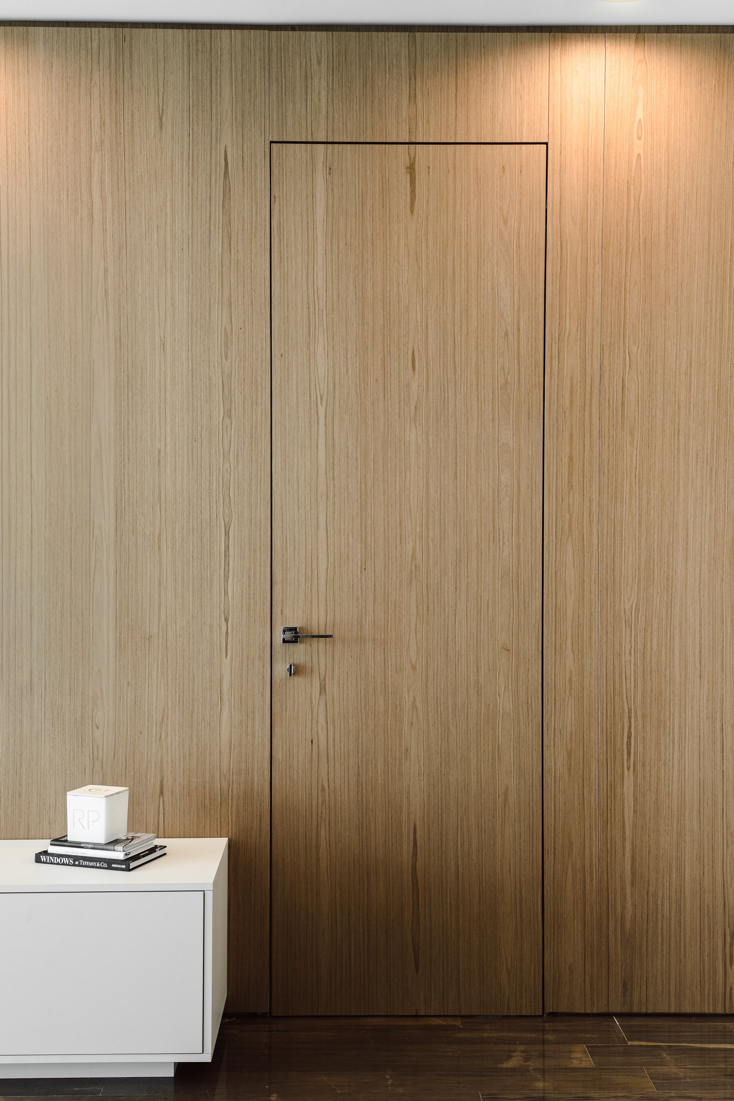 Close-up of a wooden wall with a door, and a small white table holding a stack of books and a white cube with 'RP' on it. The books include titles related to Windows and Tiffany & Co.