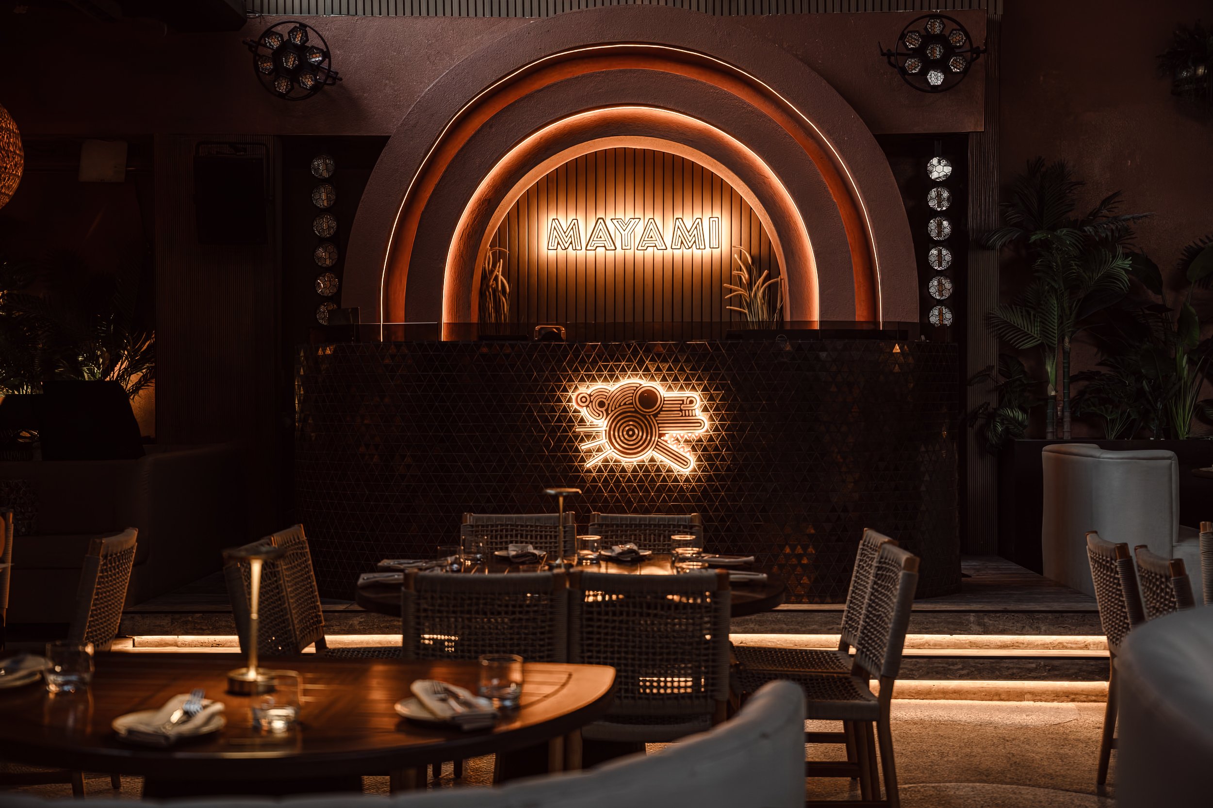 Interior of a restaurant with a illuminated sign reading 'MAYAMI' on a wooden backdrop, surrounded by plants and decorative wall lighting, with tables and chairs in the foreground.