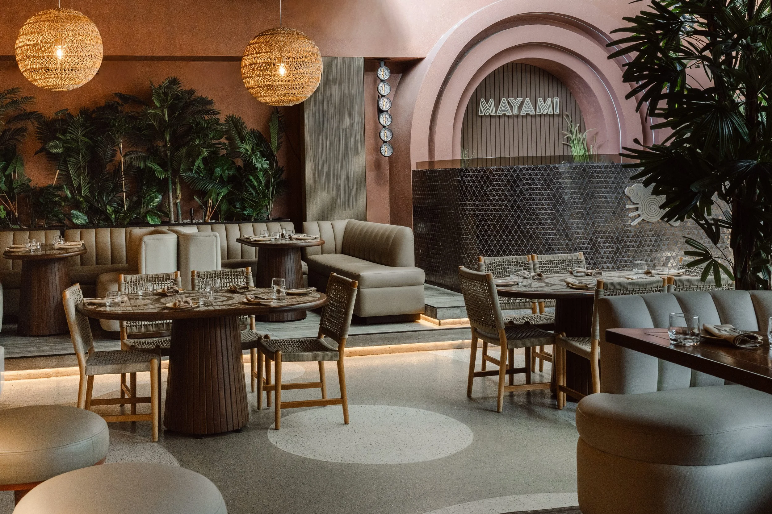 Elegant restaurant interior with round tables, woven chairs, plush seating, tropical plants, decorative lighting, and a sign reading 'MAYAMI' on a textured wall.