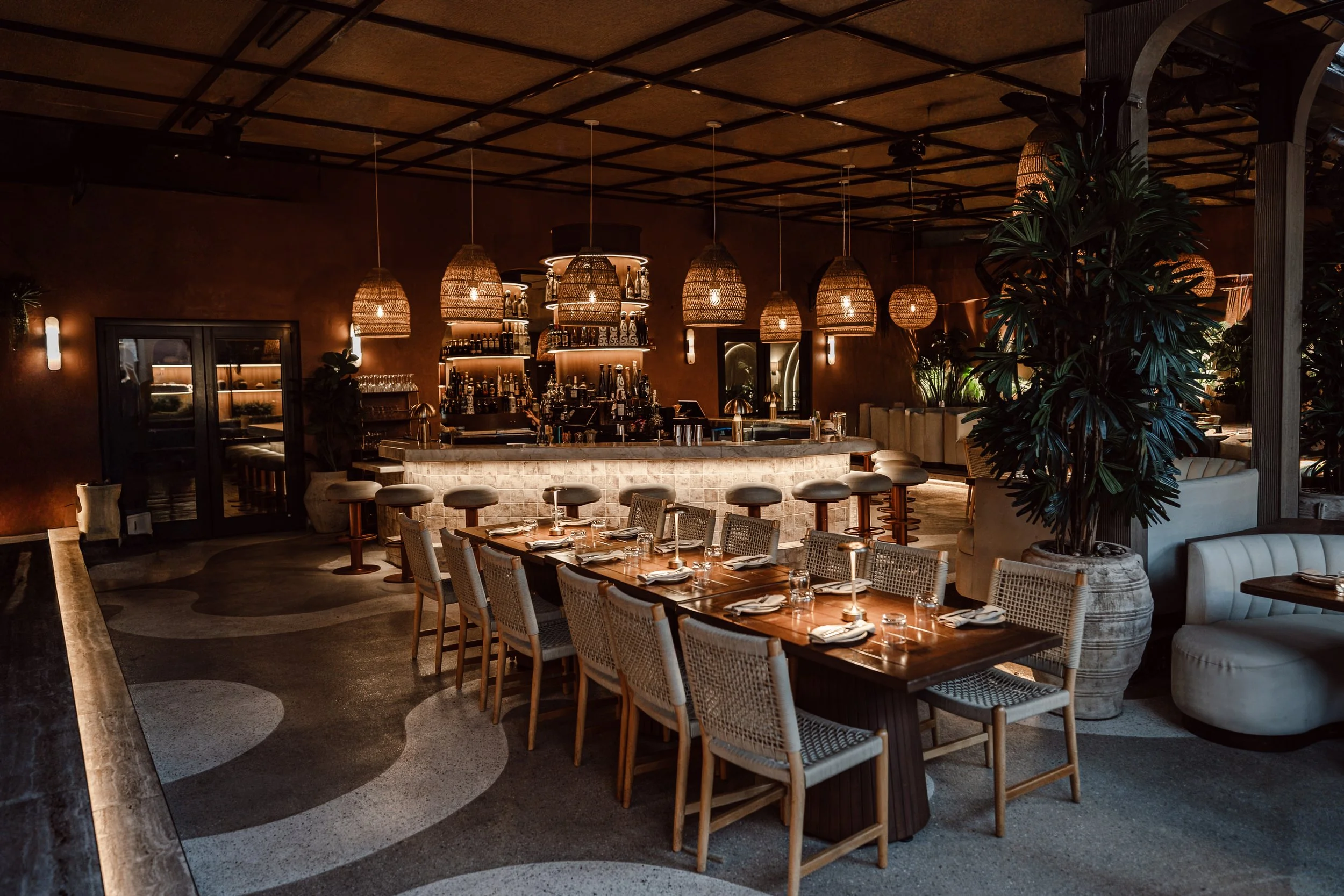 Interior of a restaurant with a bar, hanging wicker pendant lights, wooden furniture, and large potted plants.