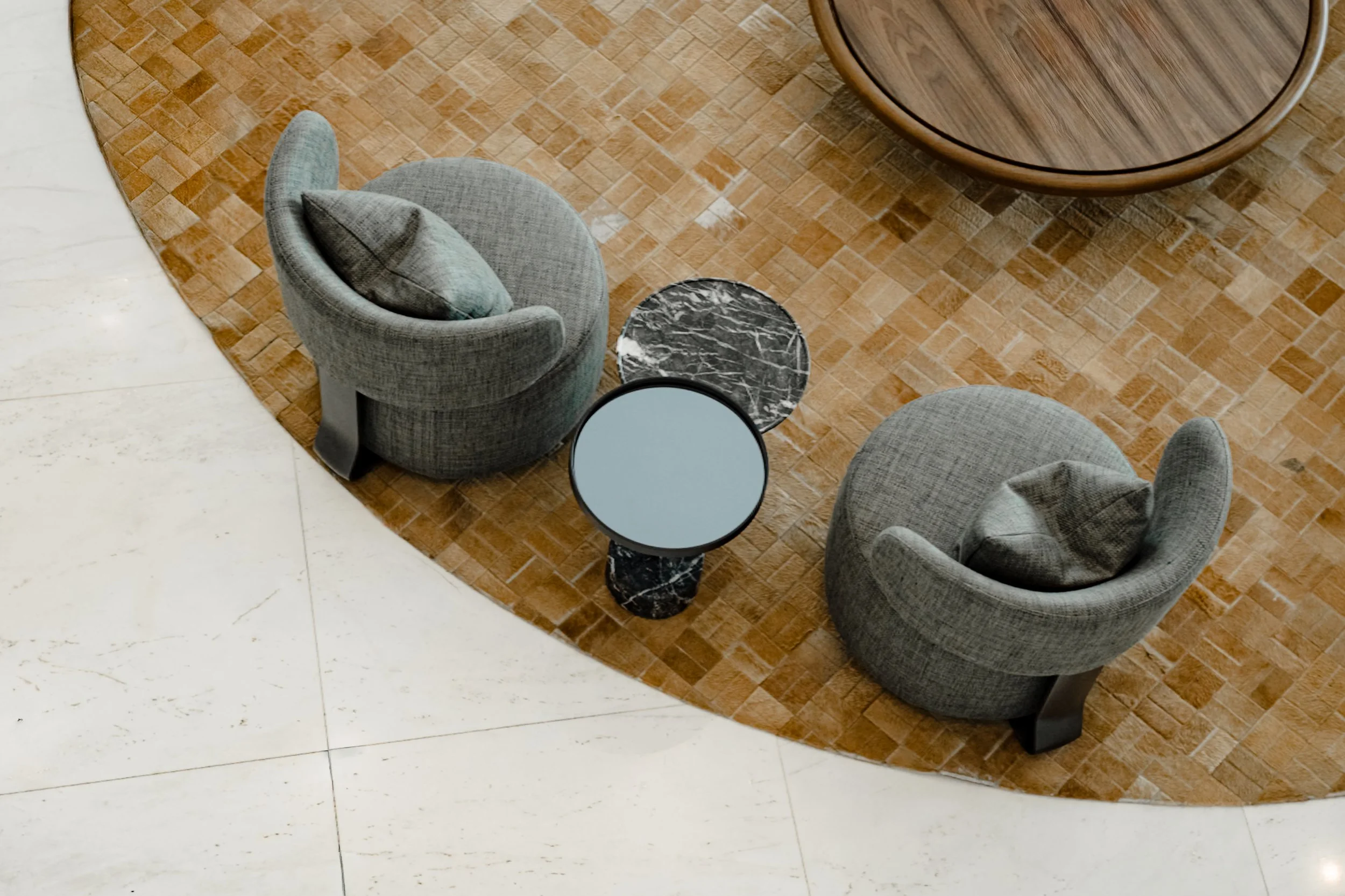 Top-down view of a sitting area with two gray upholstered armchairs, two small round coffee tables, and a wooden round table on a patterned brown rug, with white tiled flooring visible around.