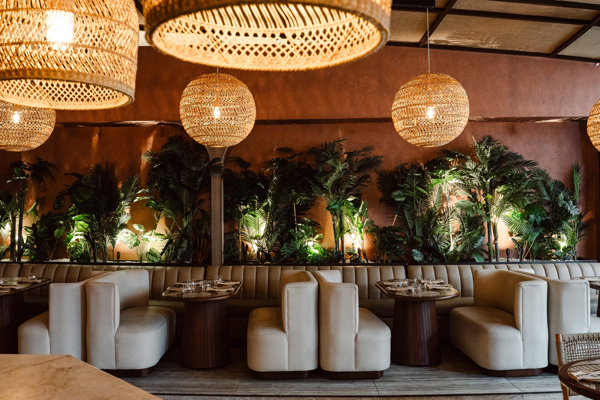 Interior of a restaurant with white cushioned seating, wooden tables, lush green plants on the wall, and woven pendant lights hanging from the ceiling.