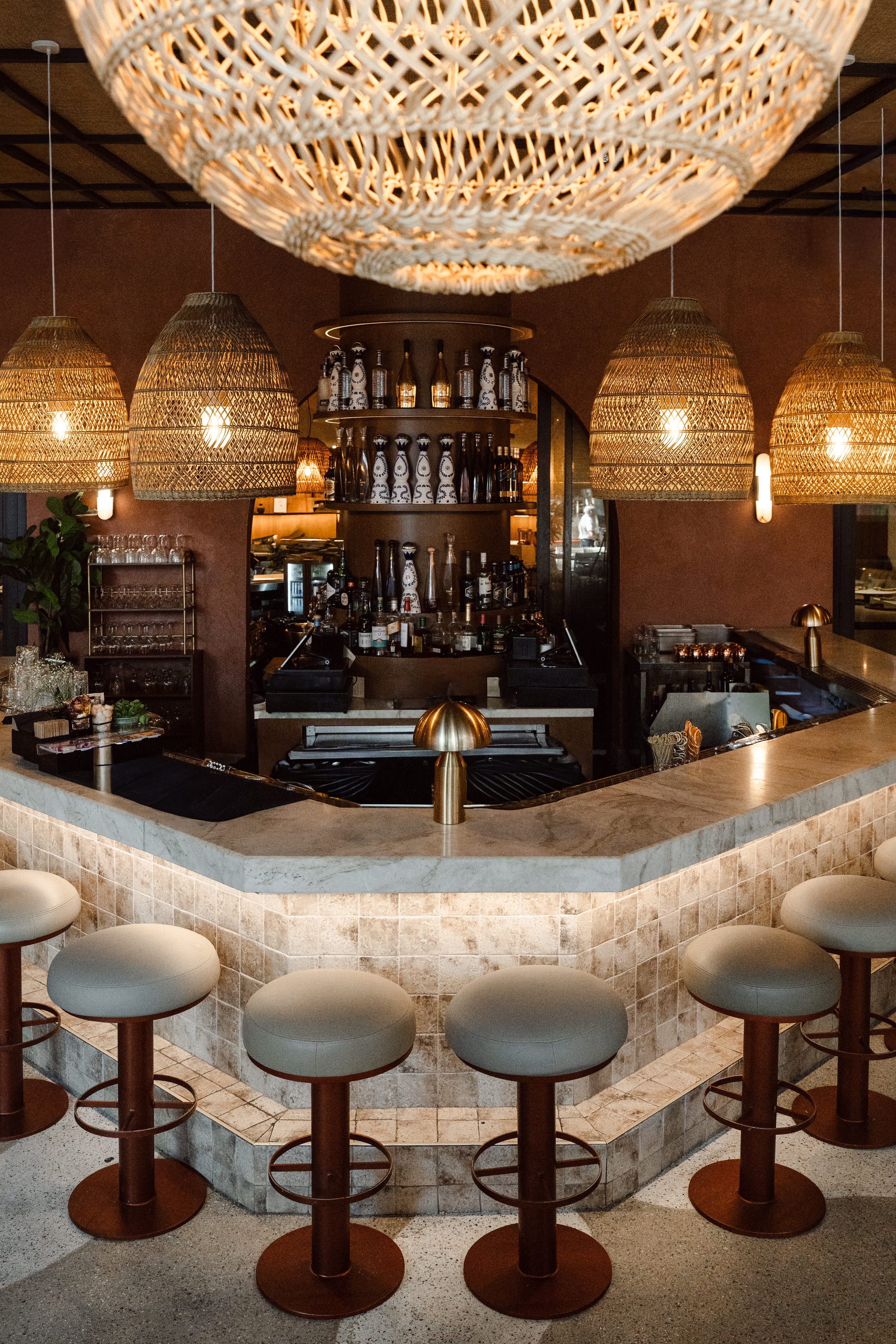 A bar counter with white bar stools around it, wicker pendant lights hanging from the ceiling, and bottles of alcohol on shelves in the background.