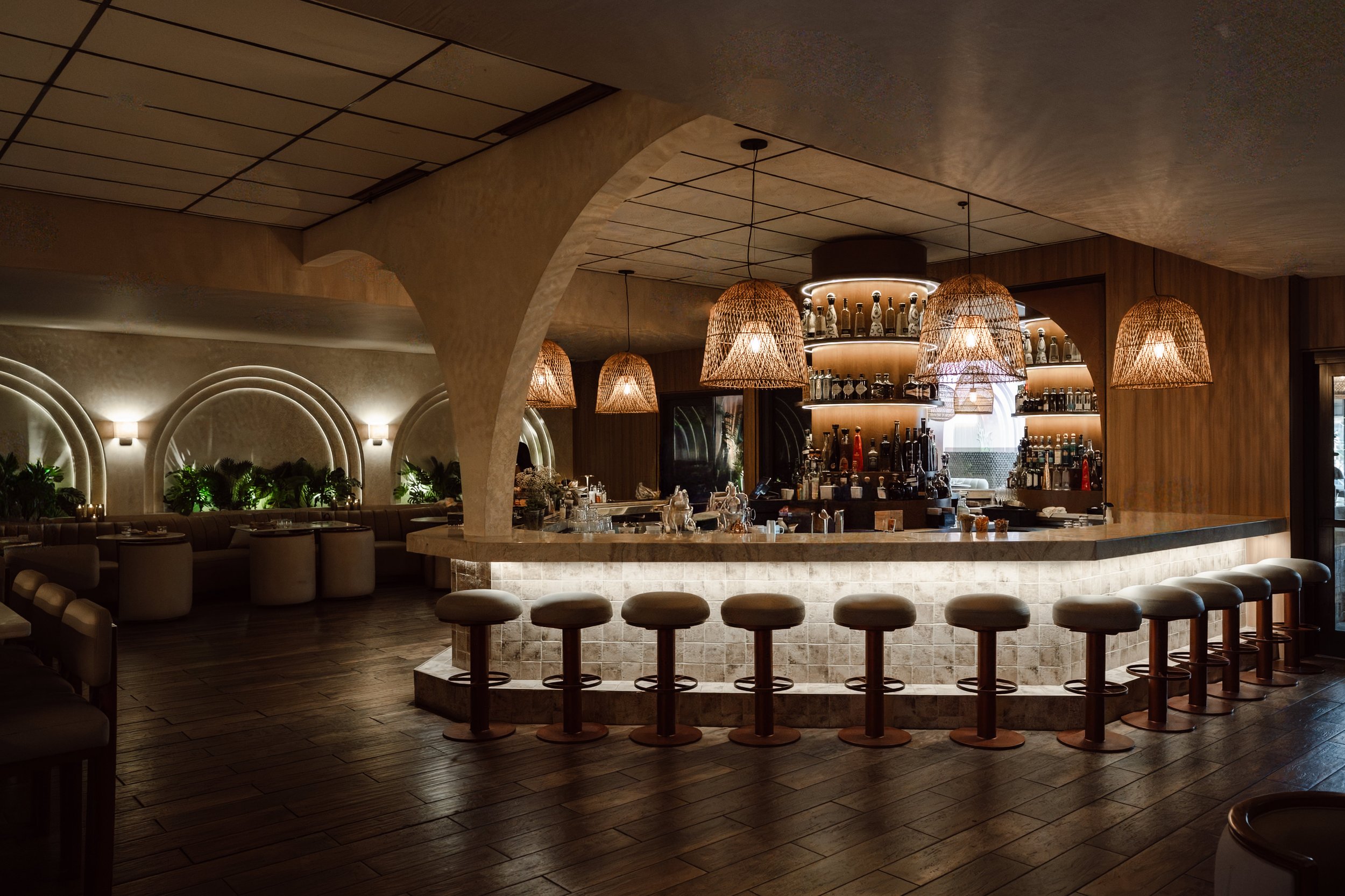 A modern restaurant interior with a circular bar area, hanging wicker pendant lights, and a wooden ceiling and walls, illuminated with warm lighting.