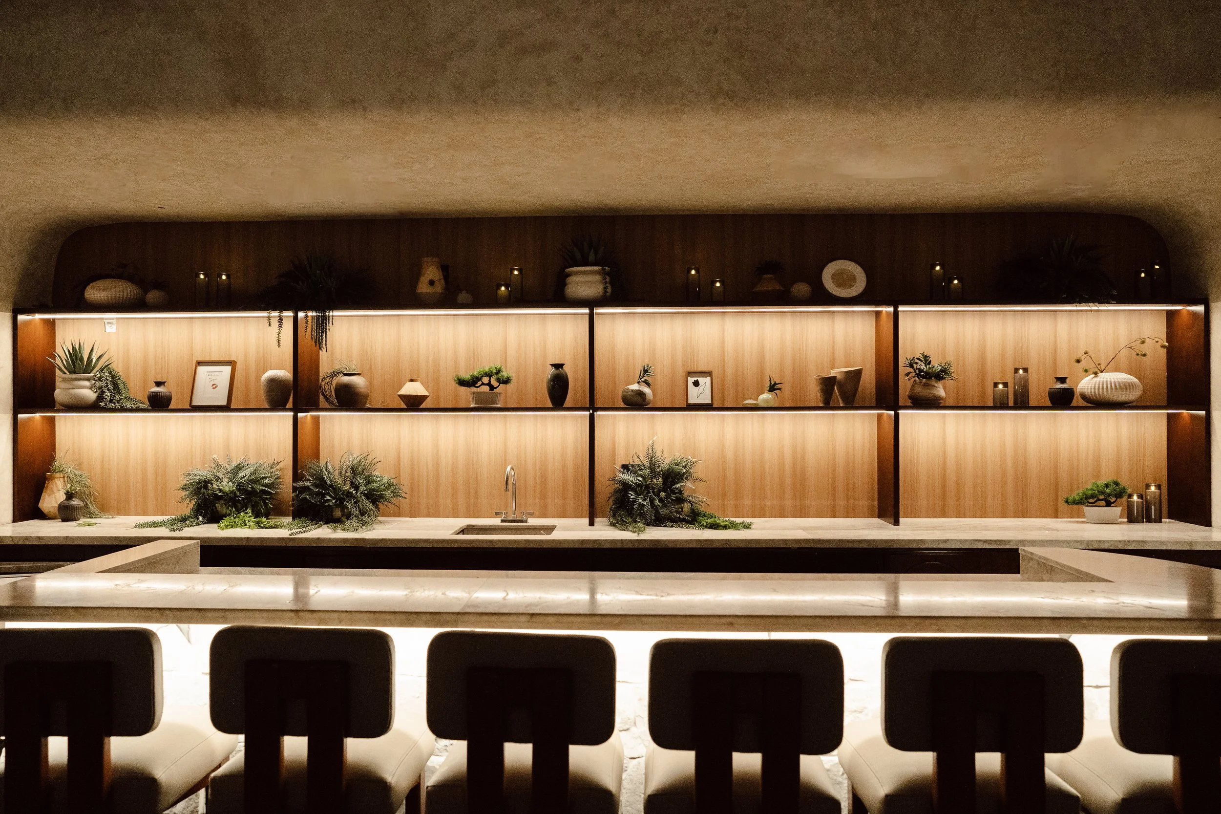 Modern kitchen with wooden shelving displaying potted plants, decorative objects, and candles, illuminated with warm lighting.