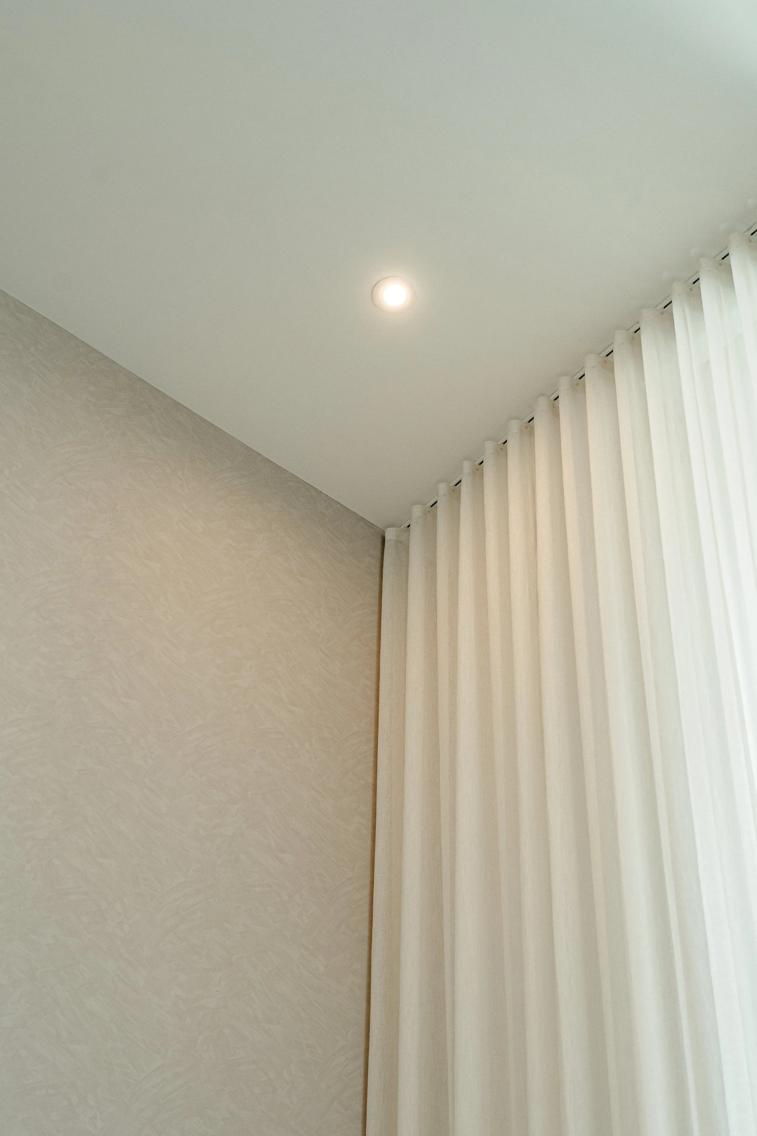 A ceiling with a single recessed light and white curtains hanging beside a window.