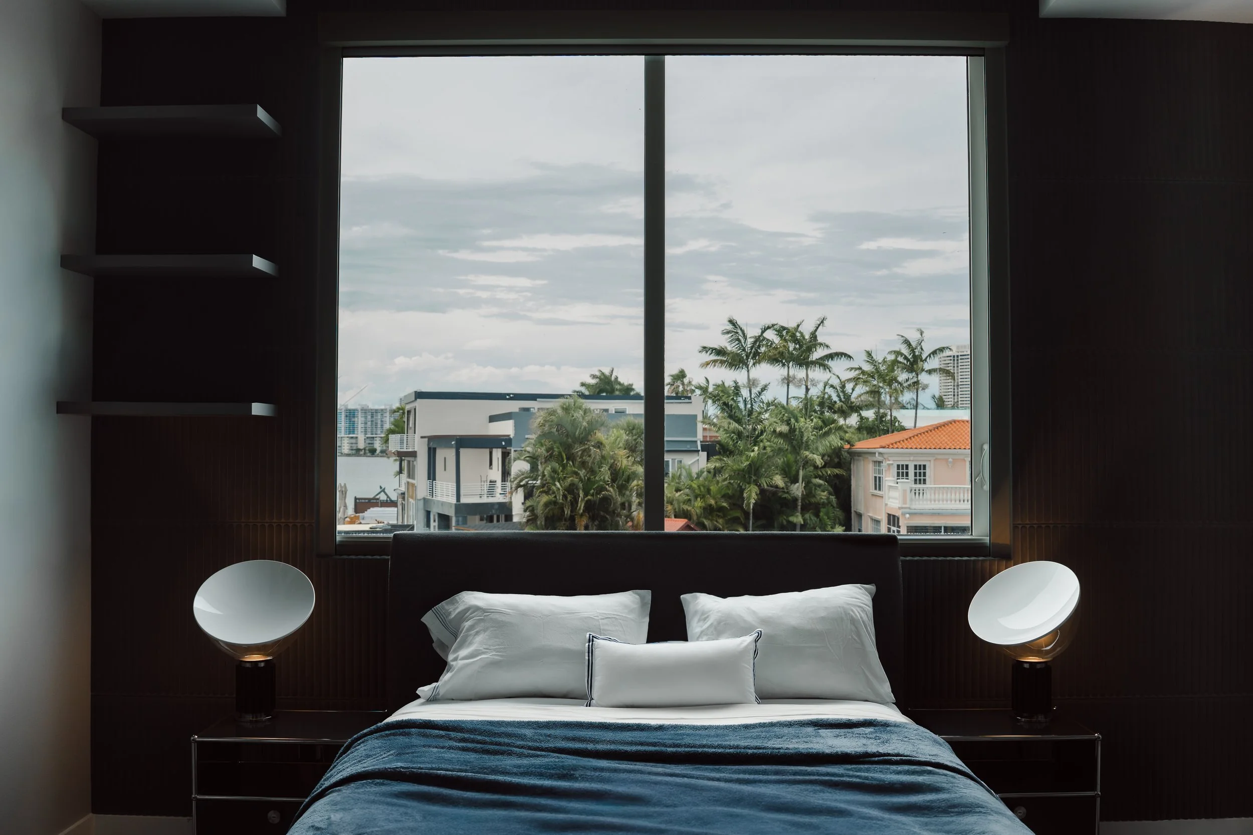 Modern bedroom with large window showing a cityscape with palm trees. The bed has white pillows and a dark blue blanket, flanked by two black bedside lamps with white shades.