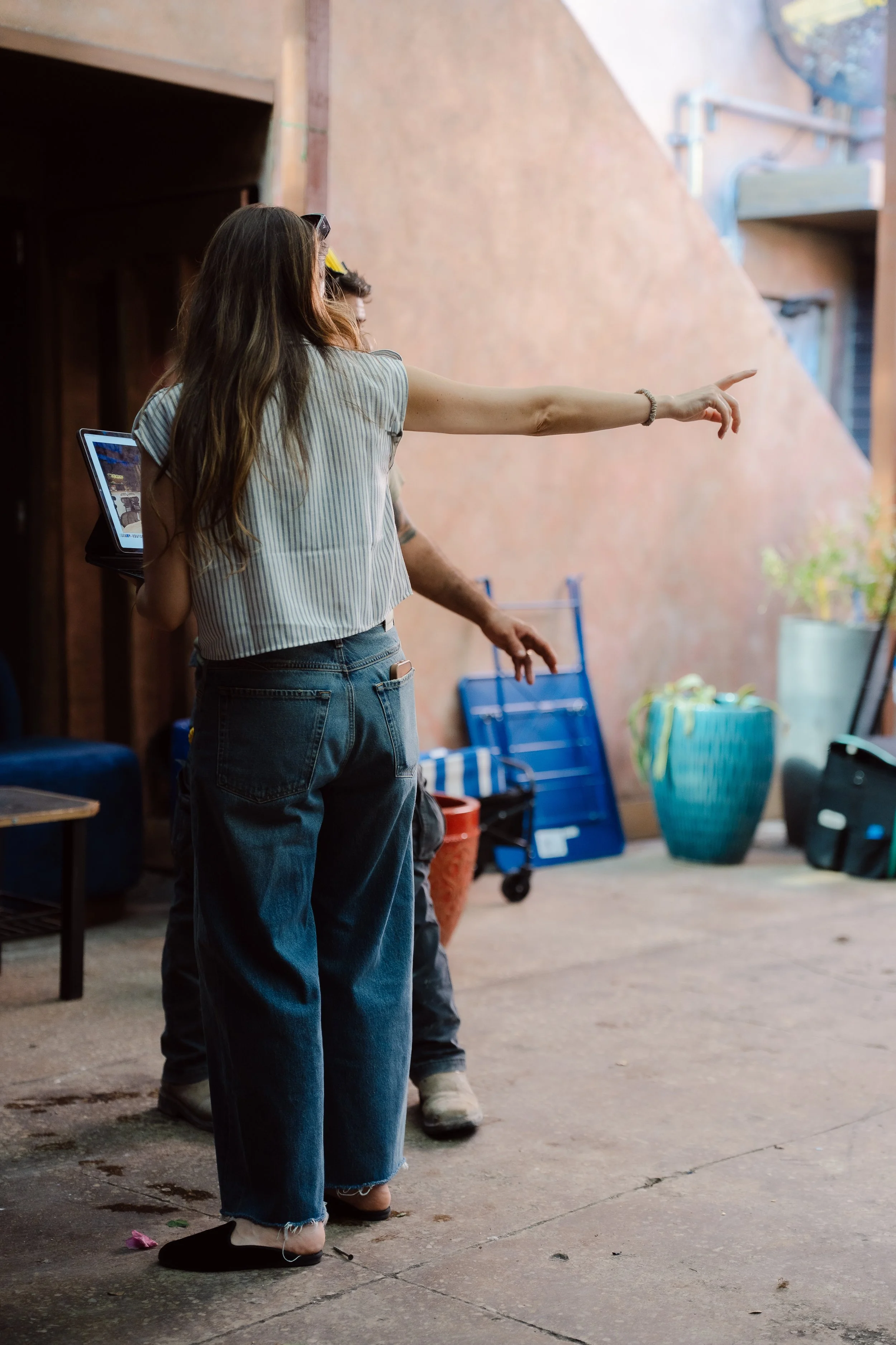 Three people standing in a semi-circle in an indoor space, with one person pointing towards something. The people have long hair and are wearing casual clothing, including jeans and slip-on shoes. There are various objects around them, including a bl