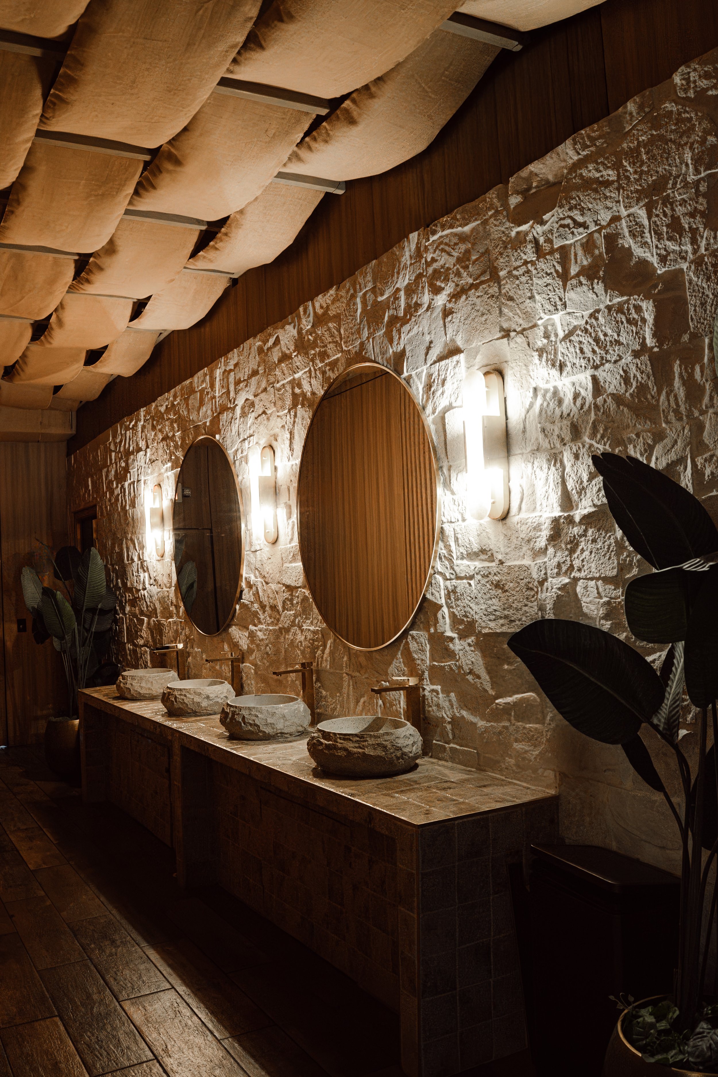 A bathroom with a rustic stone wall and wooden ceiling beams. There are three oval mirrors above a marble countertop with six stone vessel sinks. The lighting is soft and warm, creating a cozy atmosphere.