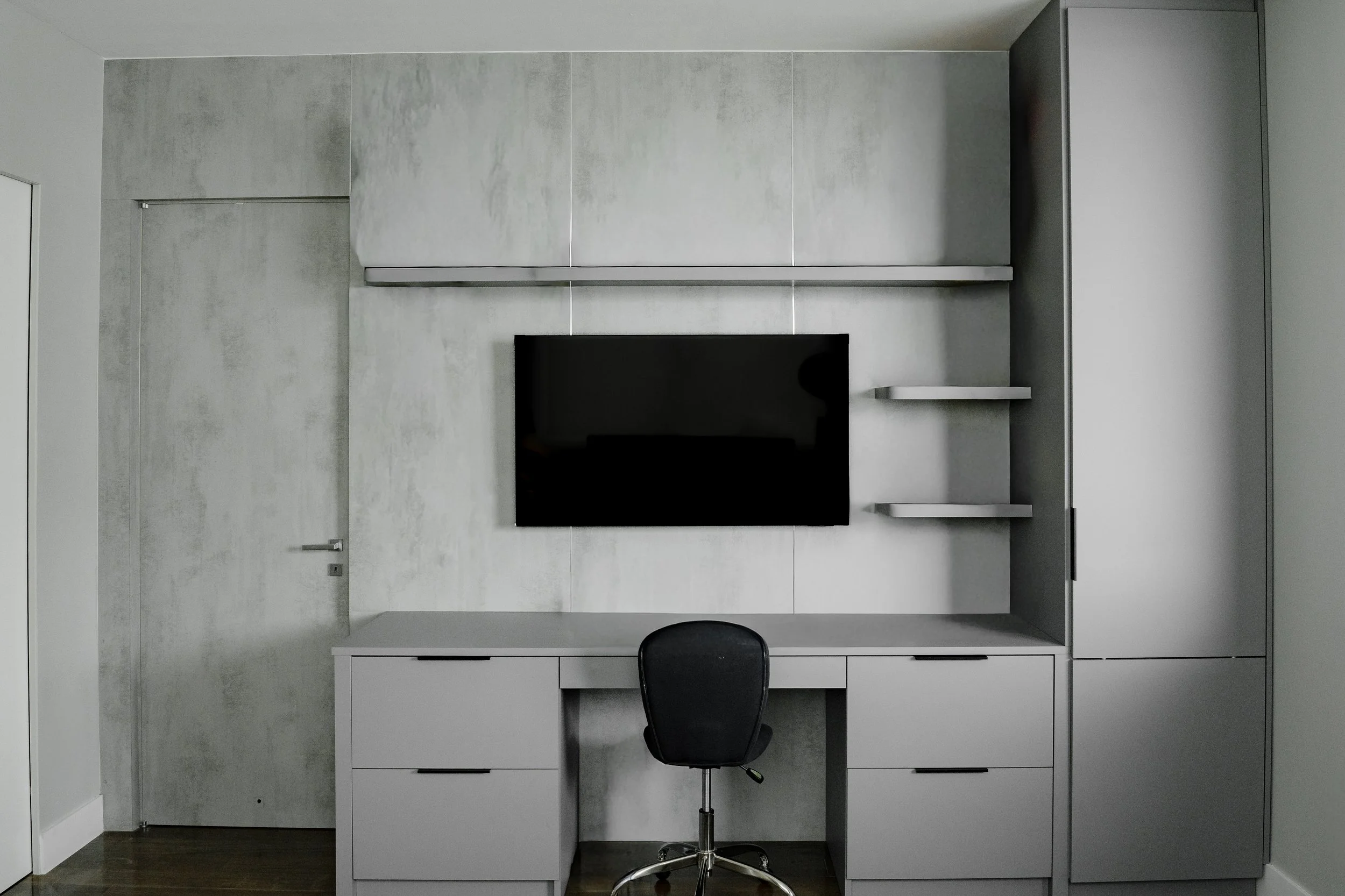 Modern gray and white built-in desk and storage unit in a small room with a black office chair and a wall-mounted flat-screen television.