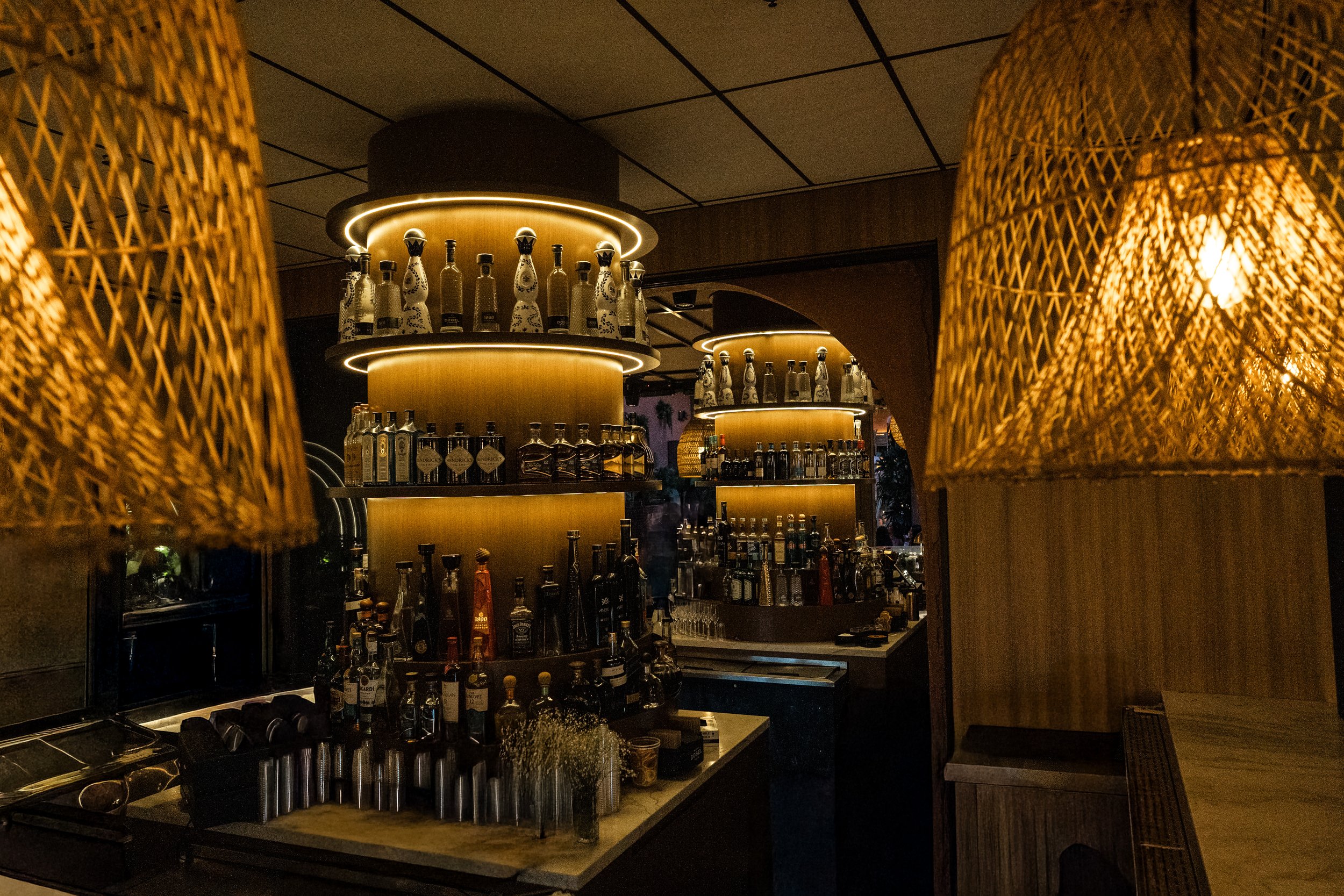 A bar interior with warm lighting, showcasing shelves filled with various bottles of alcohol, decorative woven lampshades, and a modern, cozy atmosphere.
