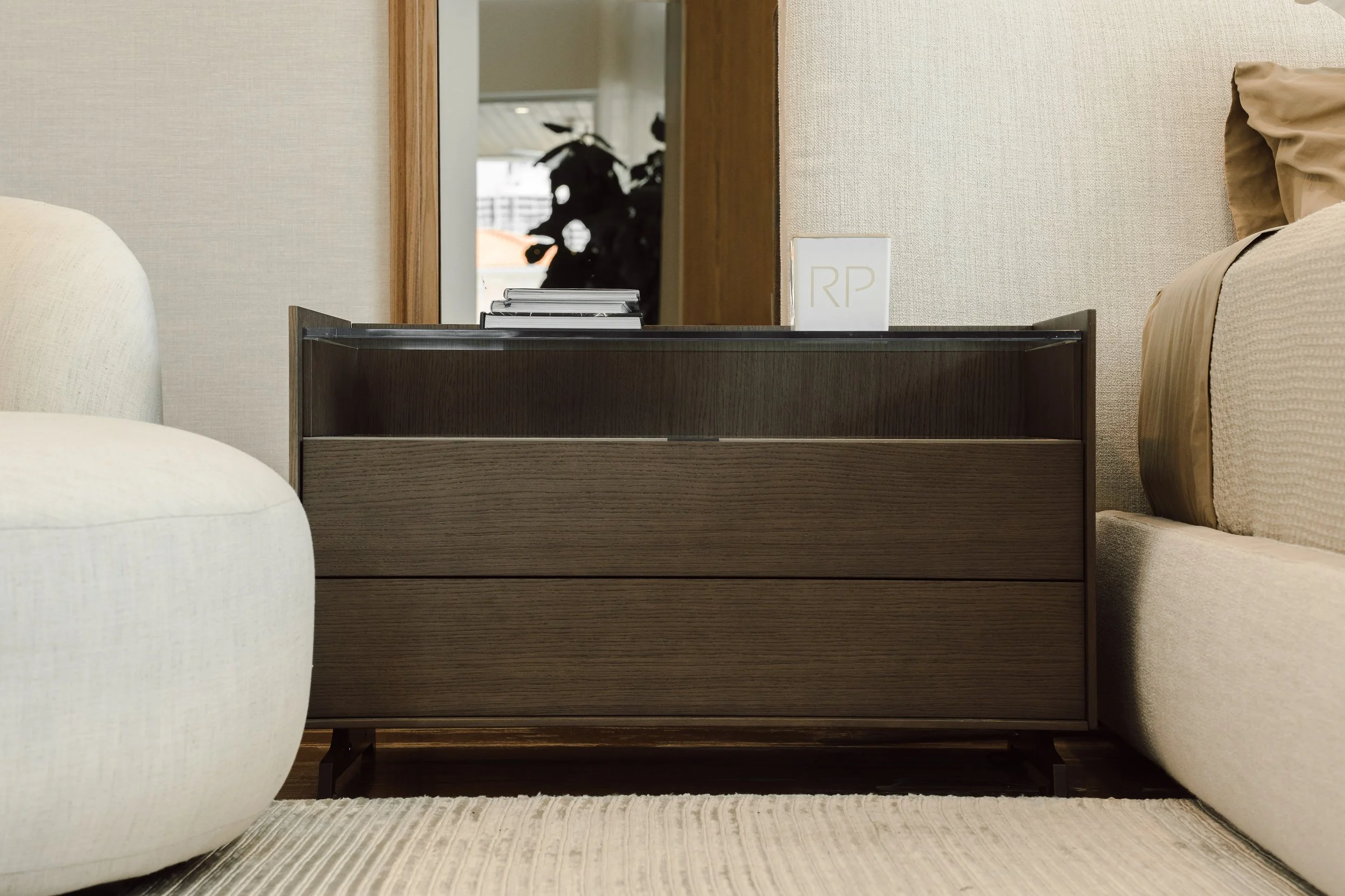 Interior of a bedroom with a dark wooden dresser, beige walls, a mirror, a white decorative object with initials 'RP', and part of a bed with a beige pillow.