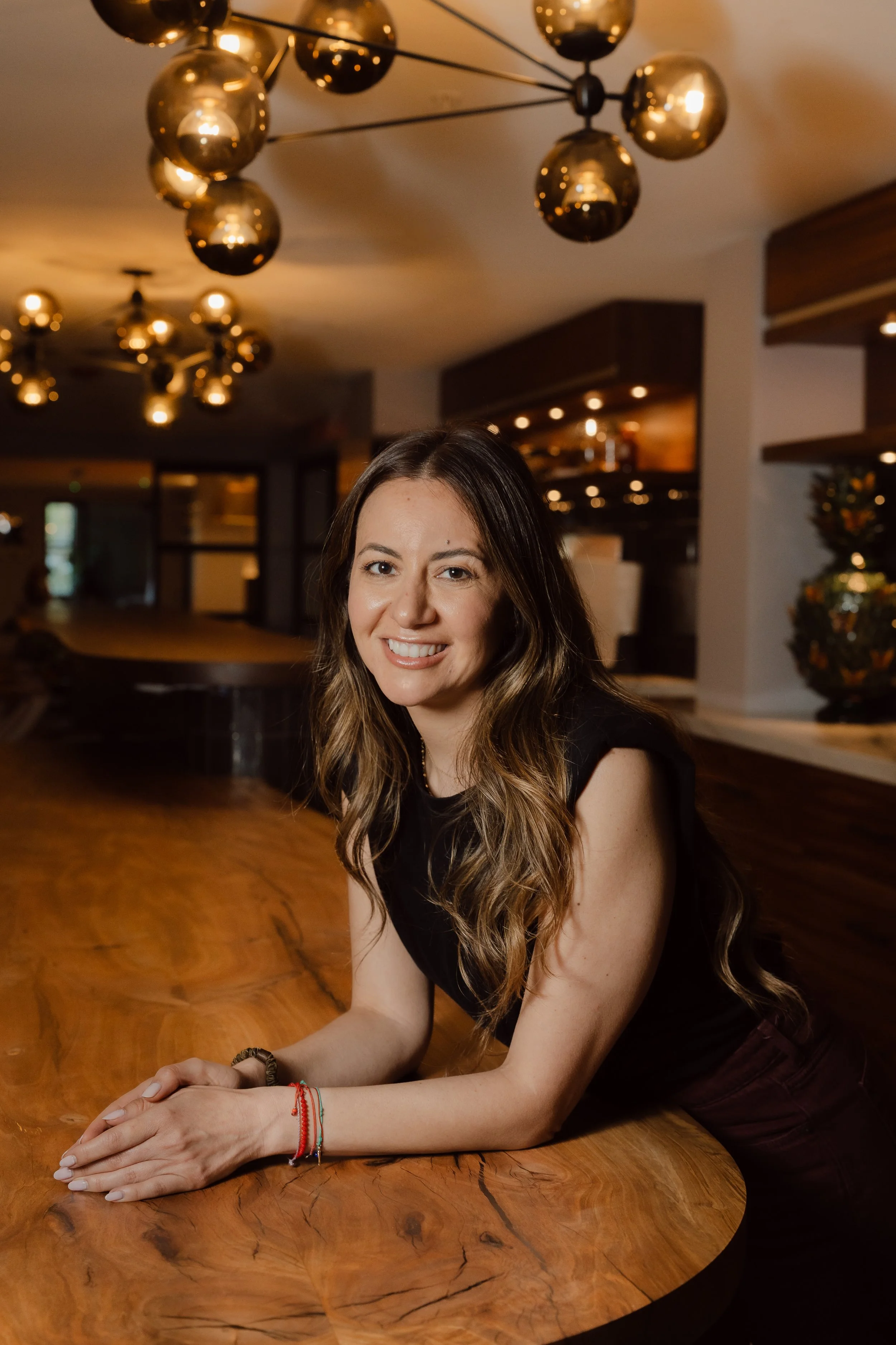 A young woman with long wavy brown hair, smiling, sitting at a polished wooden table in a warmly lit, modern interior with gold-colored spherical chandeliers hanging from the ceiling.