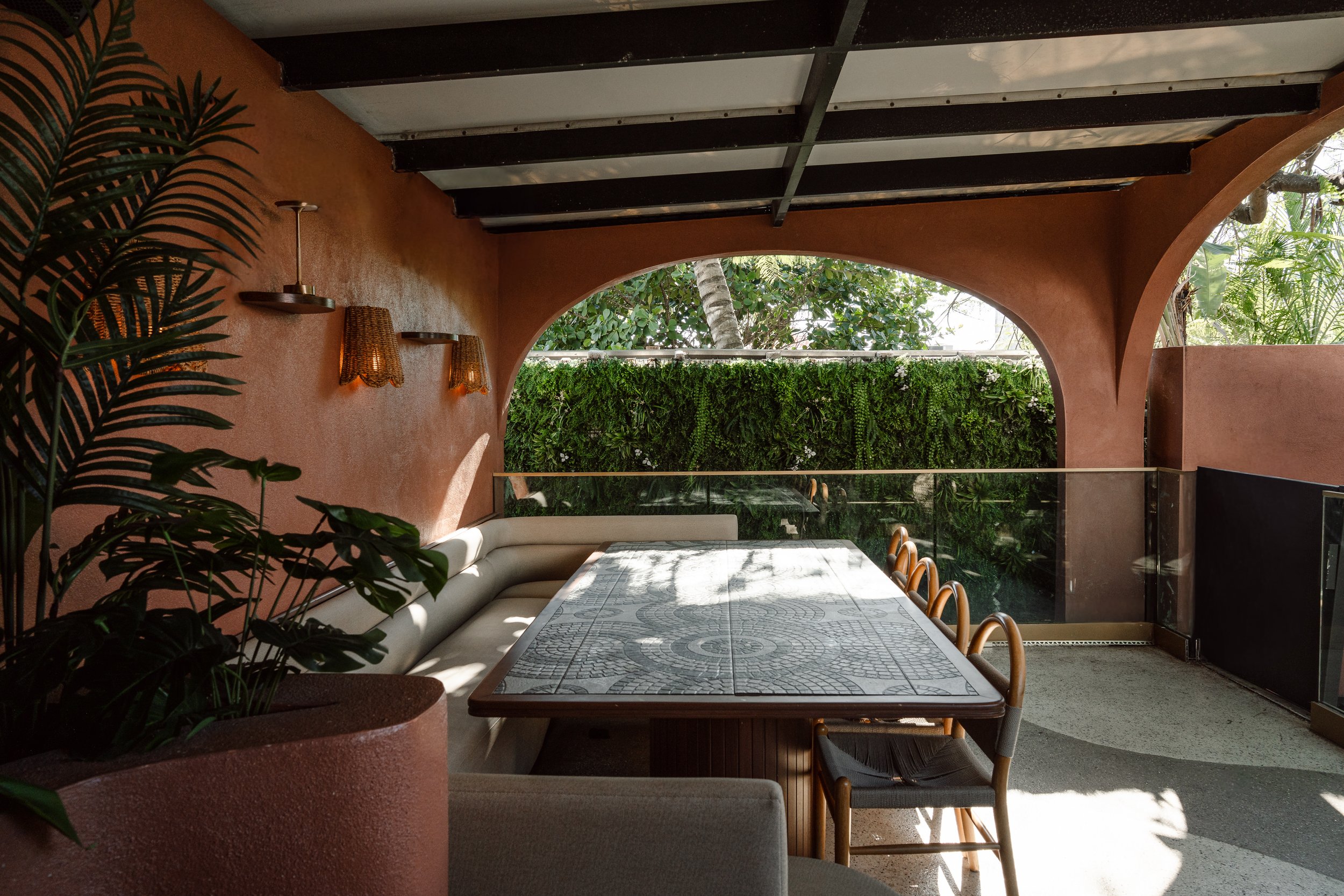 Outdoor patio area with a long rectangular dining table with a patterned surface, surrounded by chairs with light-colored cushions. The patio is partially shaded with sunlight casting shadows, and features a lush green vertical garden on one wall, an