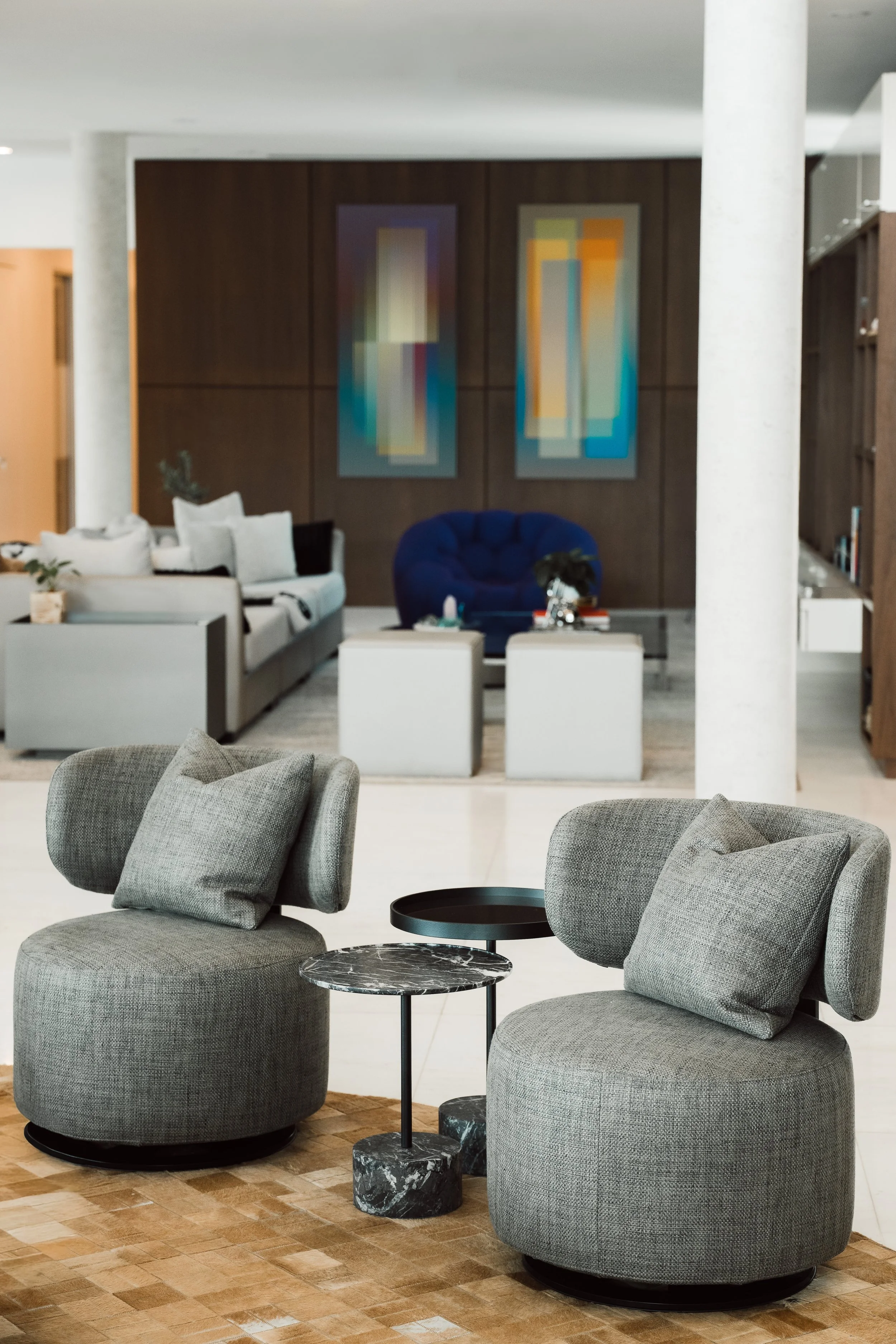 Modern living room with two gray upholstered chairs, a small round black side table, and a round marble pedestal. In the background, there is a white sofa with decorative pillows, and abstract paintings on a wood-paneled wall.