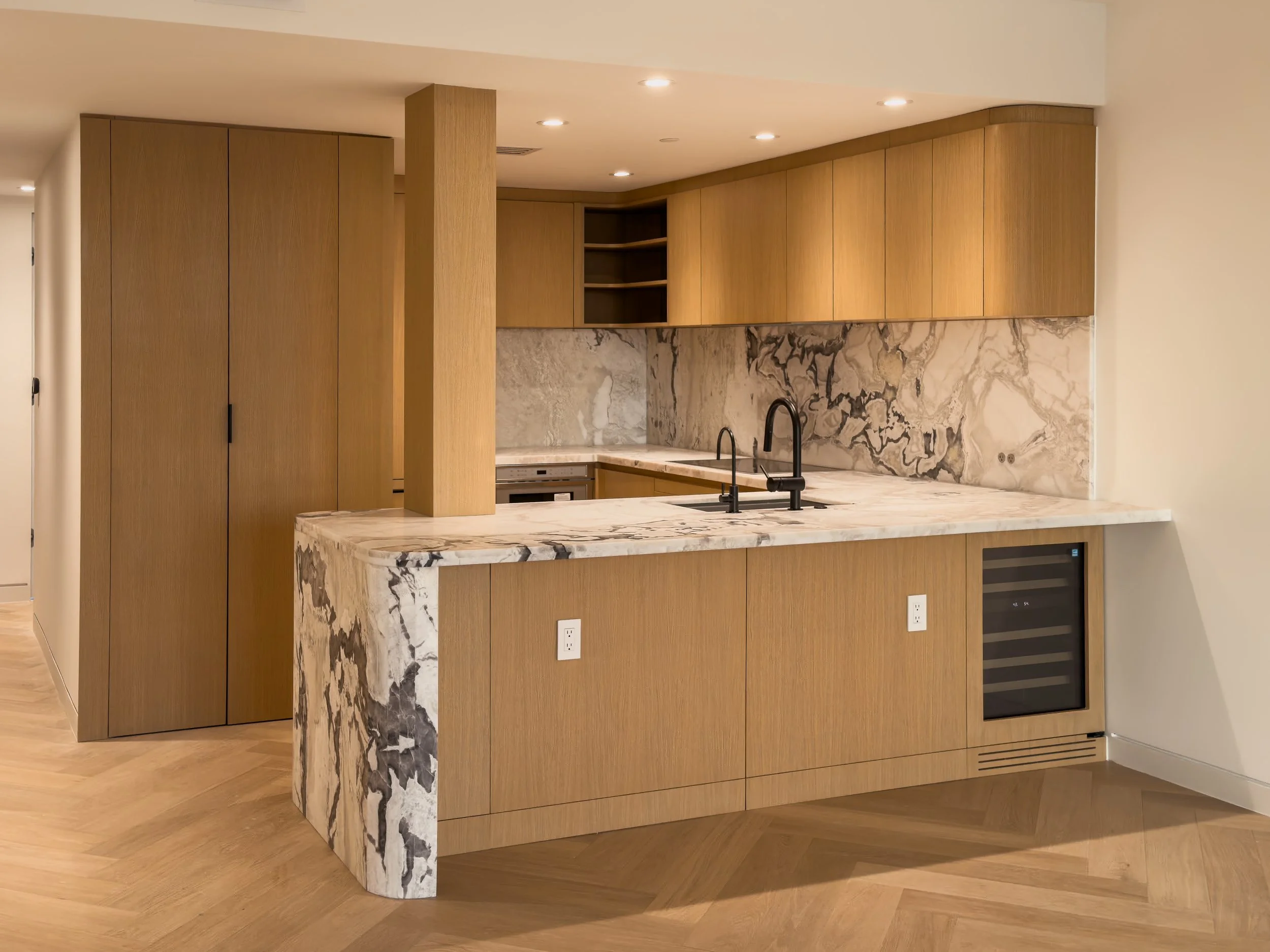 Modern kitchen with wooden cabinets, marble countertops, and a marble backsplash. Contains a black kitchen faucet, stove, and an oven. The floor has a light wood pattern and the ceiling has multiple recessed lights.