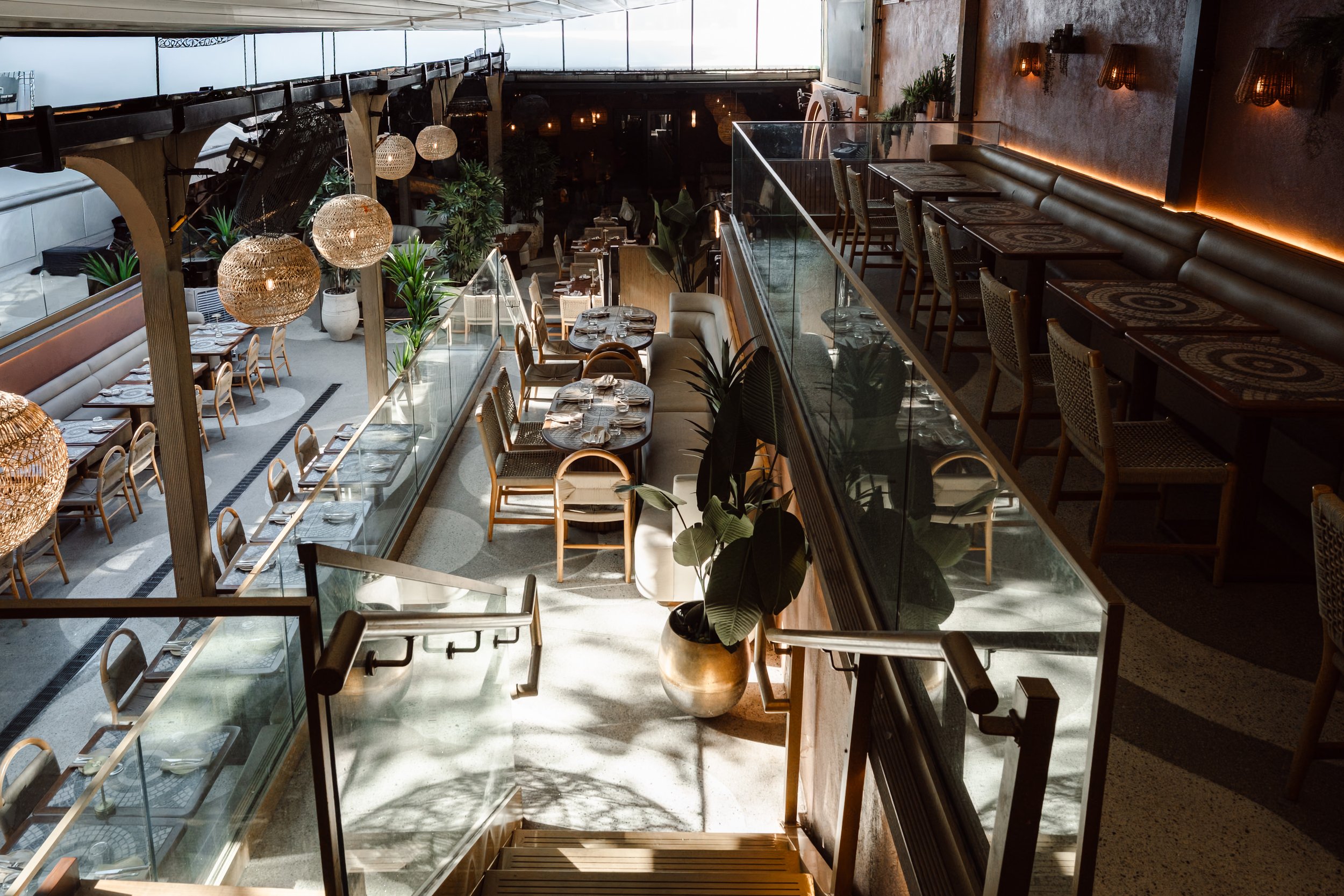 Indoor restaurant with wooden tables set with silverware and napkins, surrounded by chairs, plants, and large woven pendant lights, with a glass railing overlooking the lower level of the restaurant.