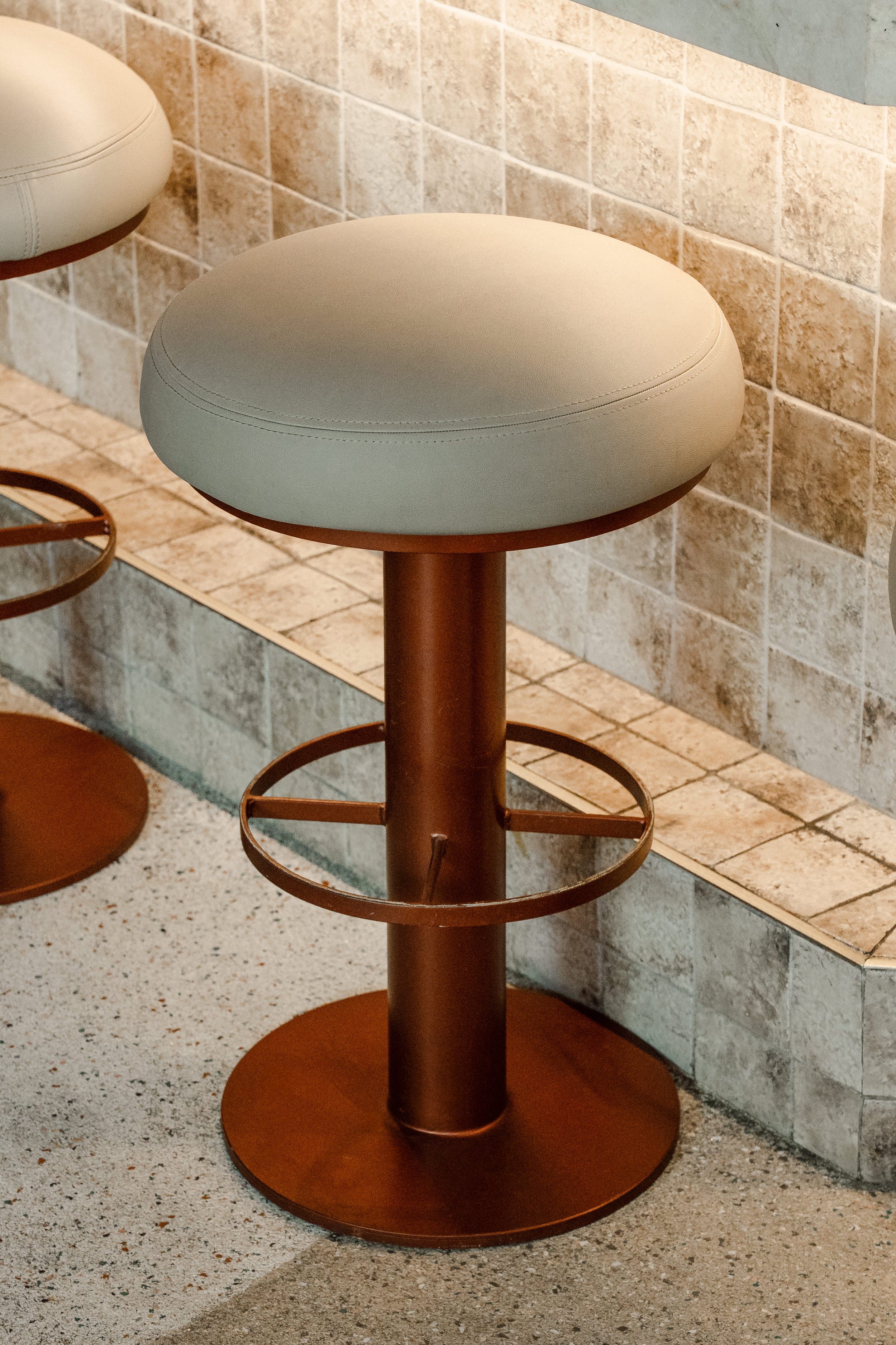 A close-up of a round beige bar stool with a brown metal base and ring support, set against a wall with beige tiles and a speckled floor.