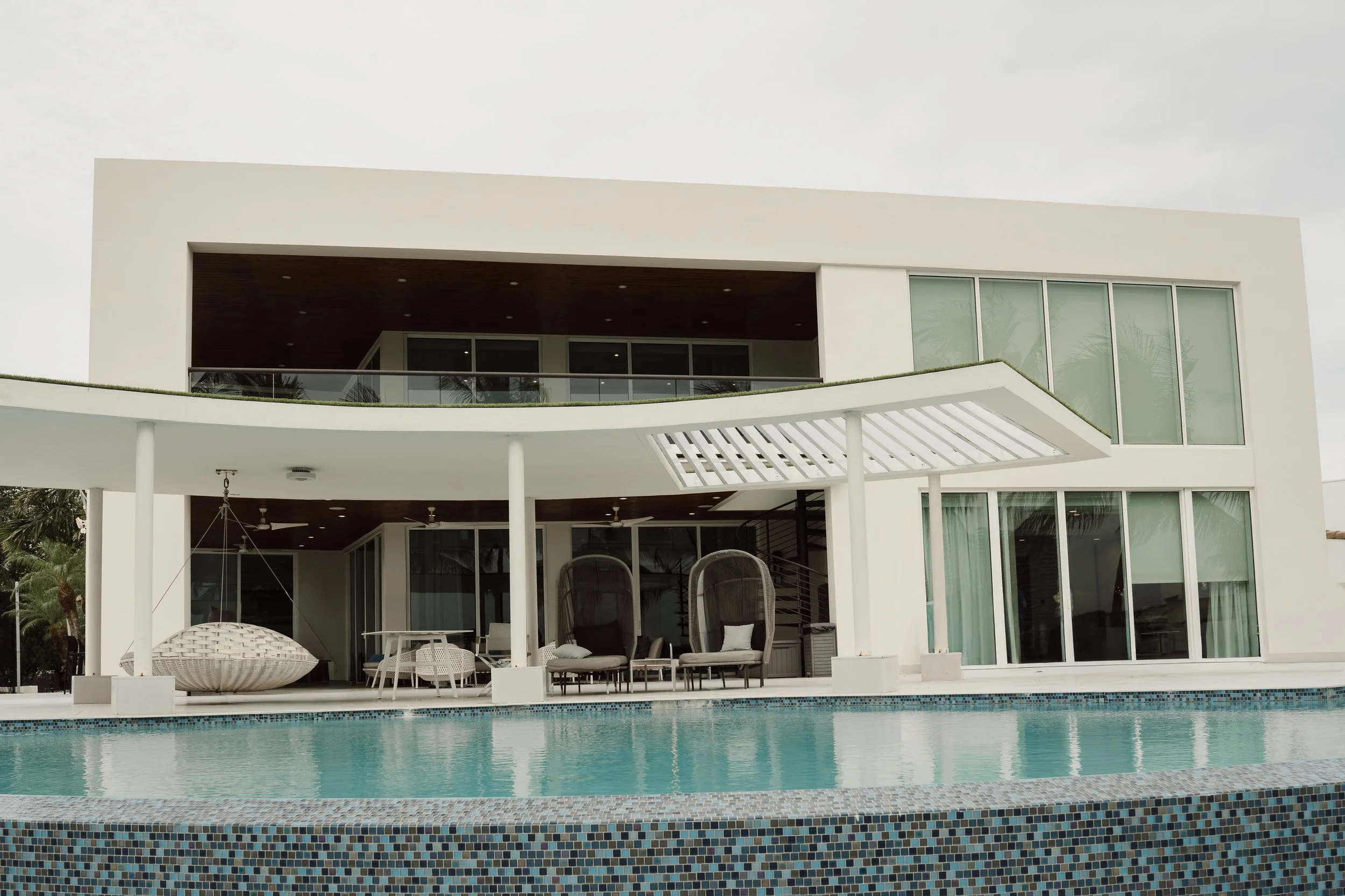 Modern white house with a pool in the front yard, outdoor furniture, and large glass windows and doors.