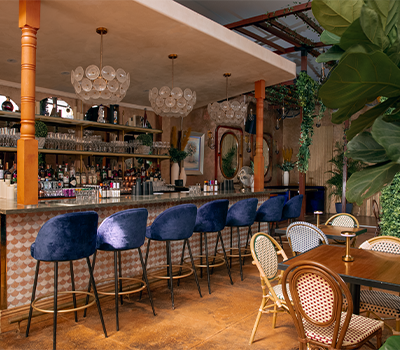 Interior of a bar with a curved marble counter, blue velvet bar stools, and a variety of bottles on shelves. Bar seating includes vintage-style chairs with cane backs, and there are large green plants and stylish lighting fixtures.
