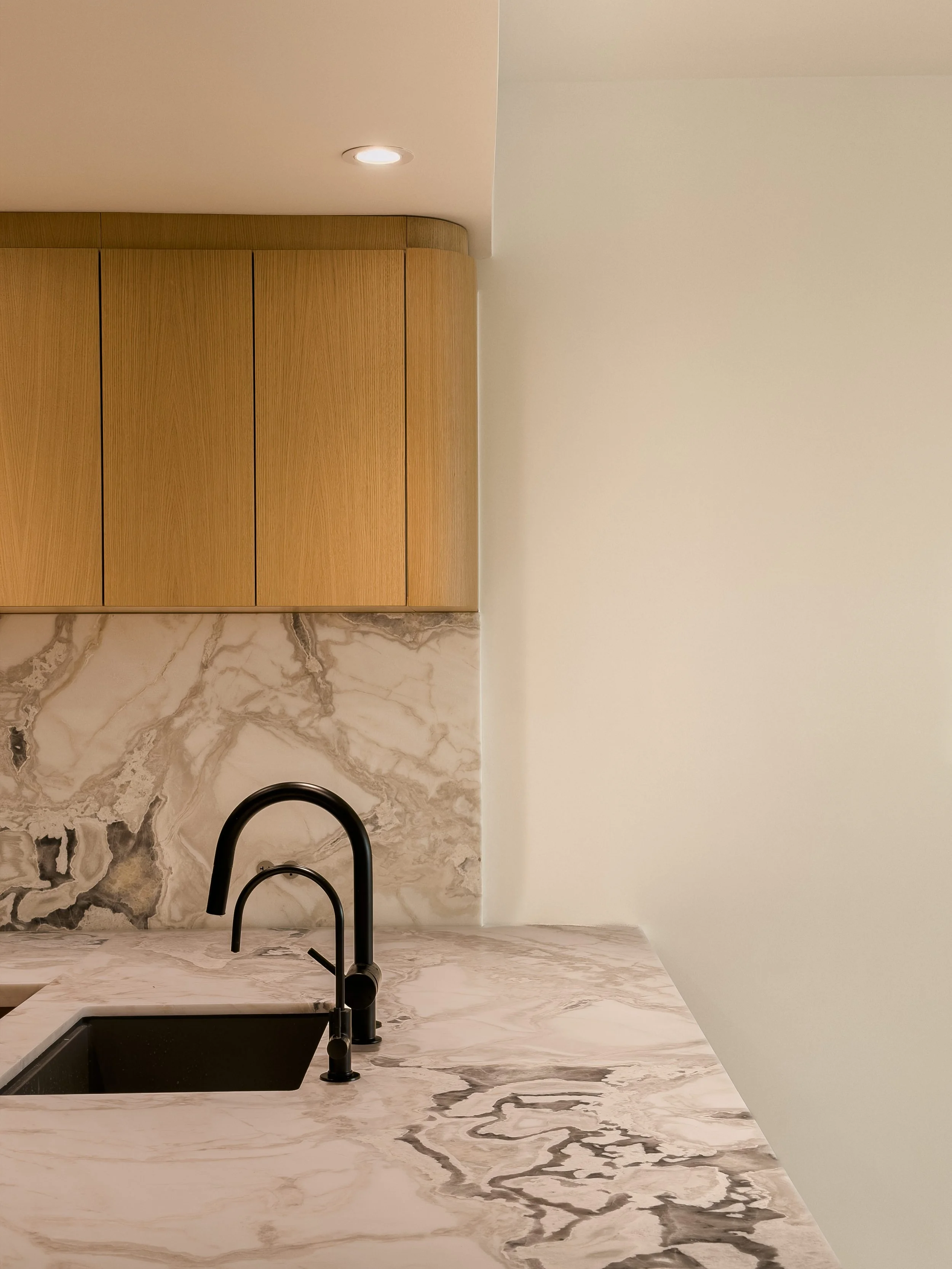 Interior kitchen with marble countertop, black sink, black faucet, wooden cabinets, and a white wall with ceiling recessed lighting.