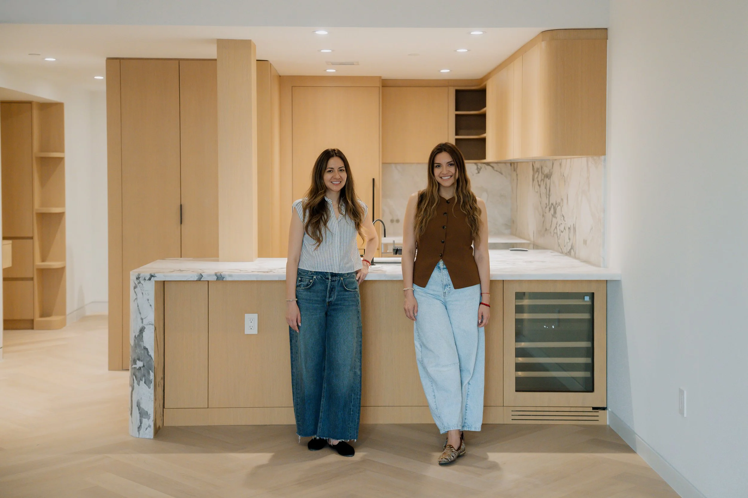 Two women smiling and standing in front of a modern kitchen with light wood cabinets and a marble countertop.
