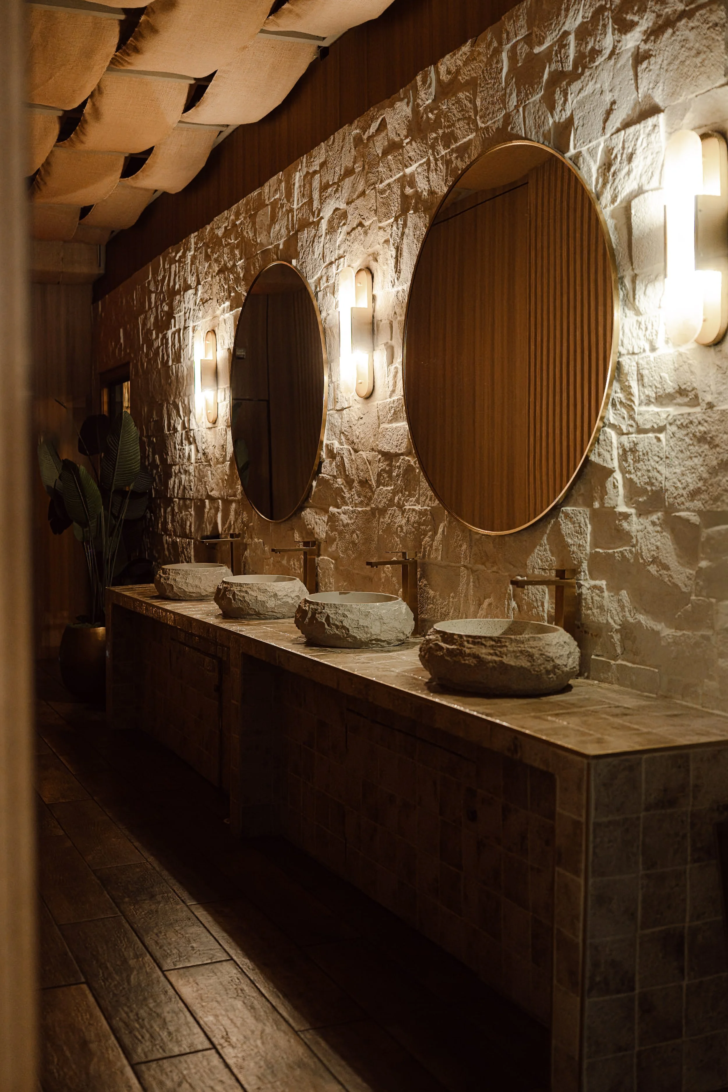 A bathroom with a stone wall, two round mirrors, and a row of rustic stone sinks on a wooden countertop, illuminated by wall-mounted lights.