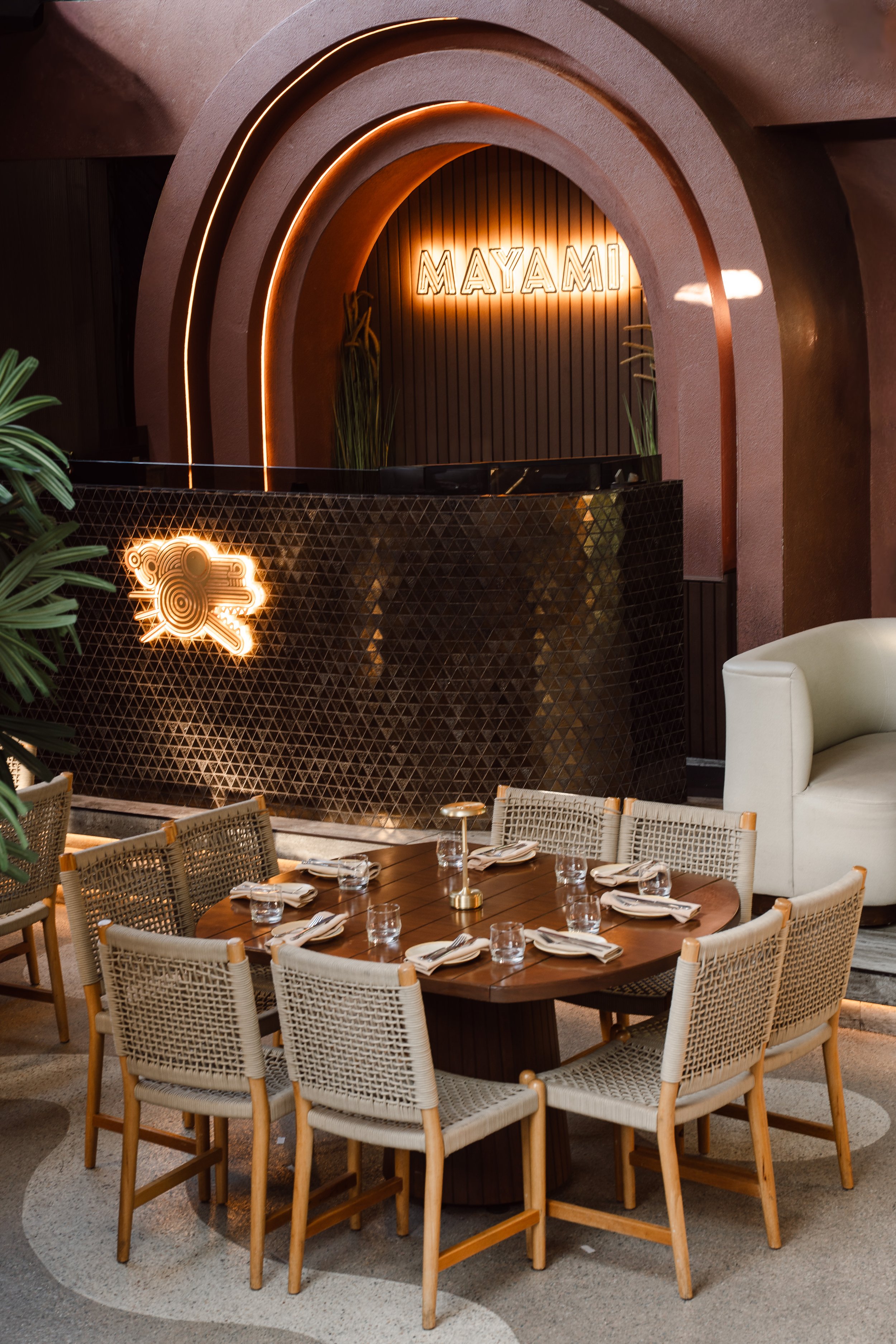 A restaurant interior with a round wooden table set for dining, surrounded by woven rattan chairs, with a festive table centerpiece and a white armchair nearby. In the background, there is a decorative wall with the word 'MAYAMI' in neon lights and a