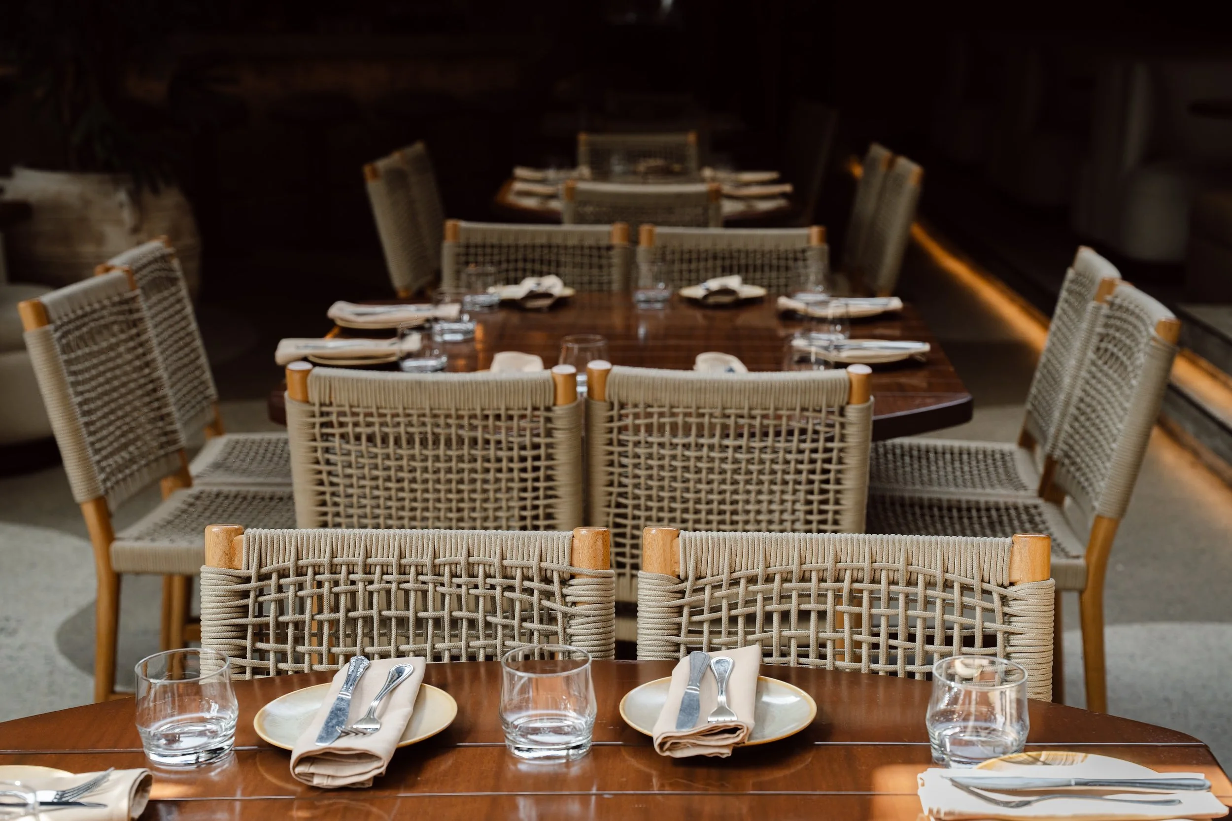 A restaurant dining area with a long wooden table set with plates, glasses, napkins, and utensils, surrounded by wicker chairs with wooden accents.