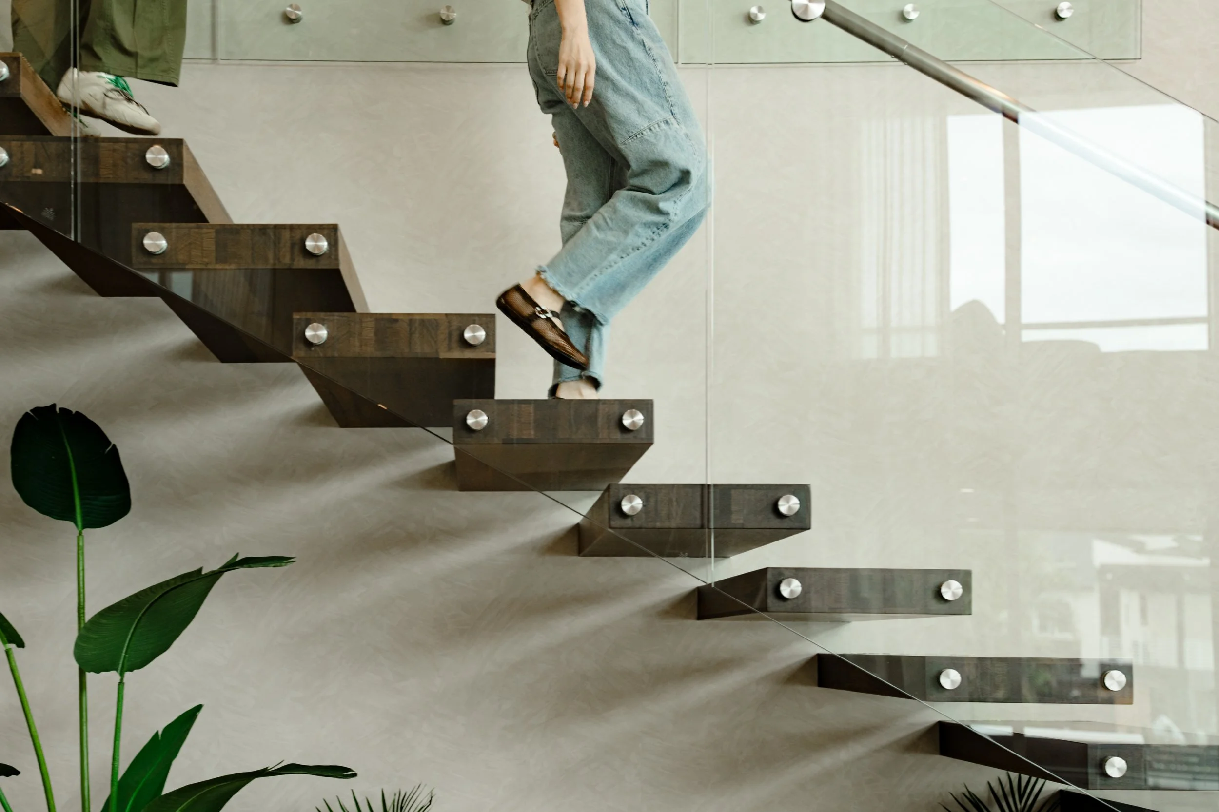 A person wearing faded jeans and black shoes stepping onto the wooden stairs of a modern staircase with glass railings, reflected in a large mirror on the wall, in a well-lit indoor space with green plants nearby.