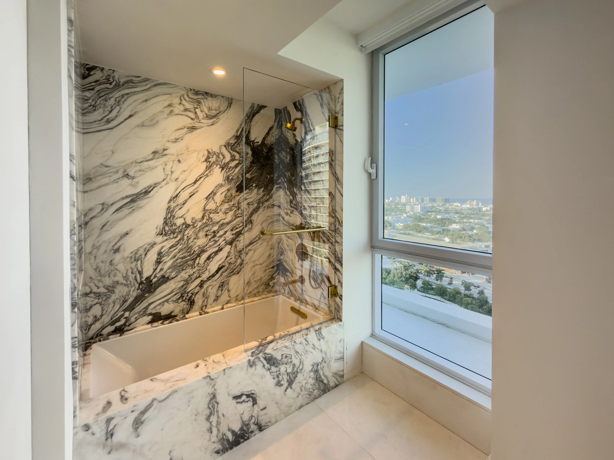 A modern bathroom shower with marble walls featuring black and white swirling patterns, a glass door, and a window showing a cityscape view with buildings and greenery.