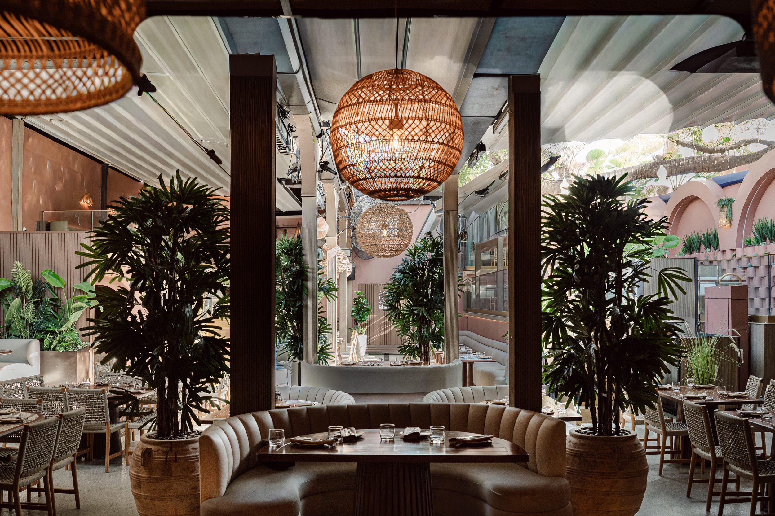 Interior of a restaurant featuring hanging wicker pendant lights, lush indoor plants, and round tables set with glasses and silverware. There is a curved booth seating area in the foreground and large windows with a view of greenery outside.