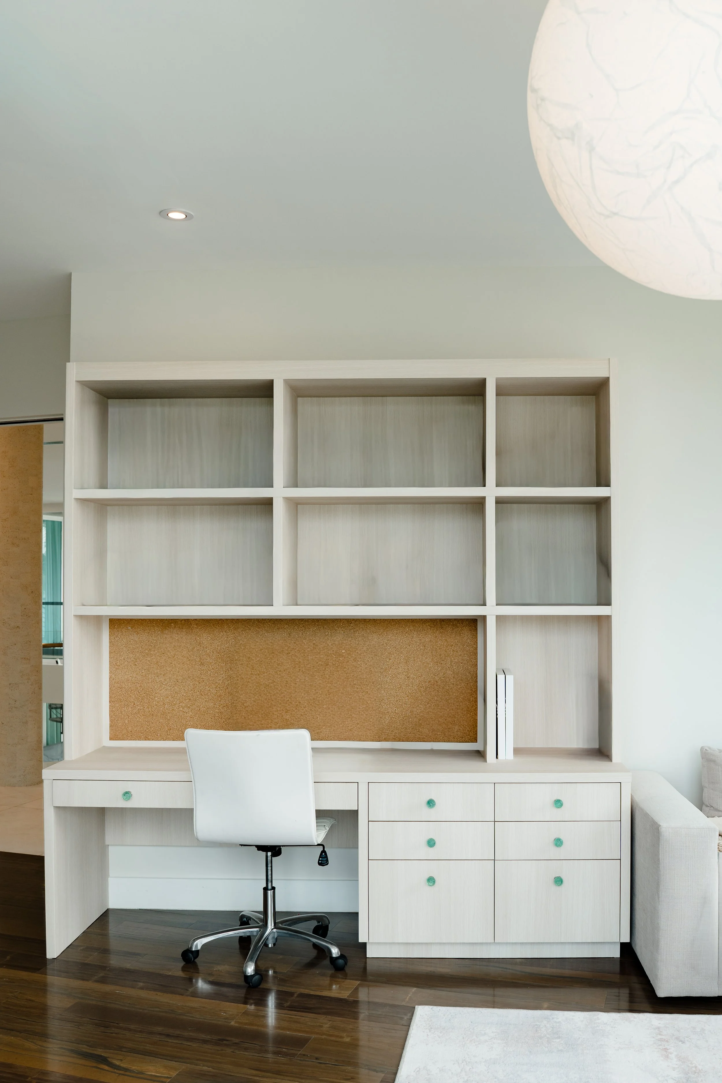 A white office desk with a swivel chair in front of a built-in bookshelf and pinboard in a modern room.