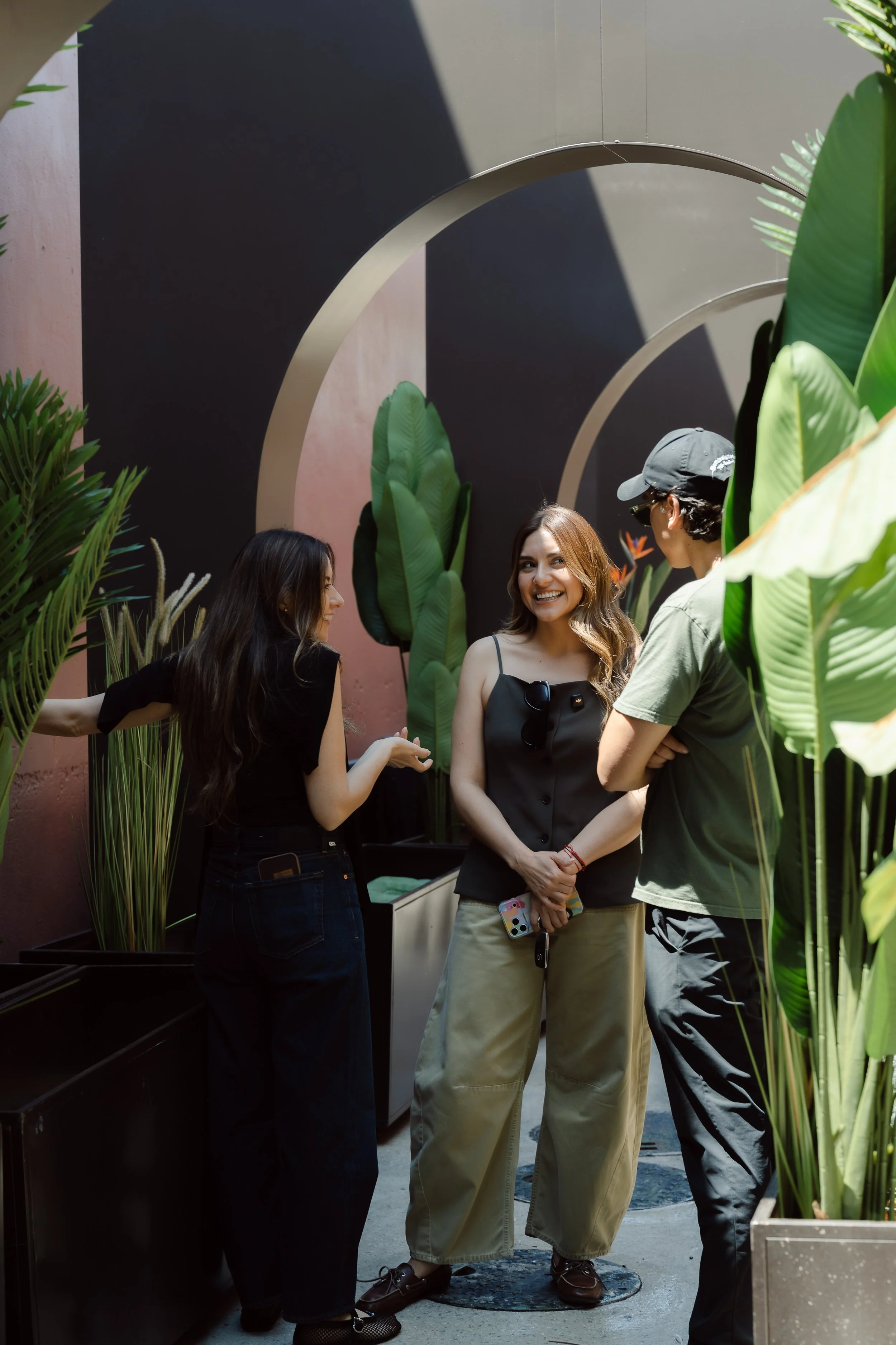 Group of four young adults standing and chatting outdoors, surrounded by large green plants, with a pink and dark gray wall with an arch design in the background.