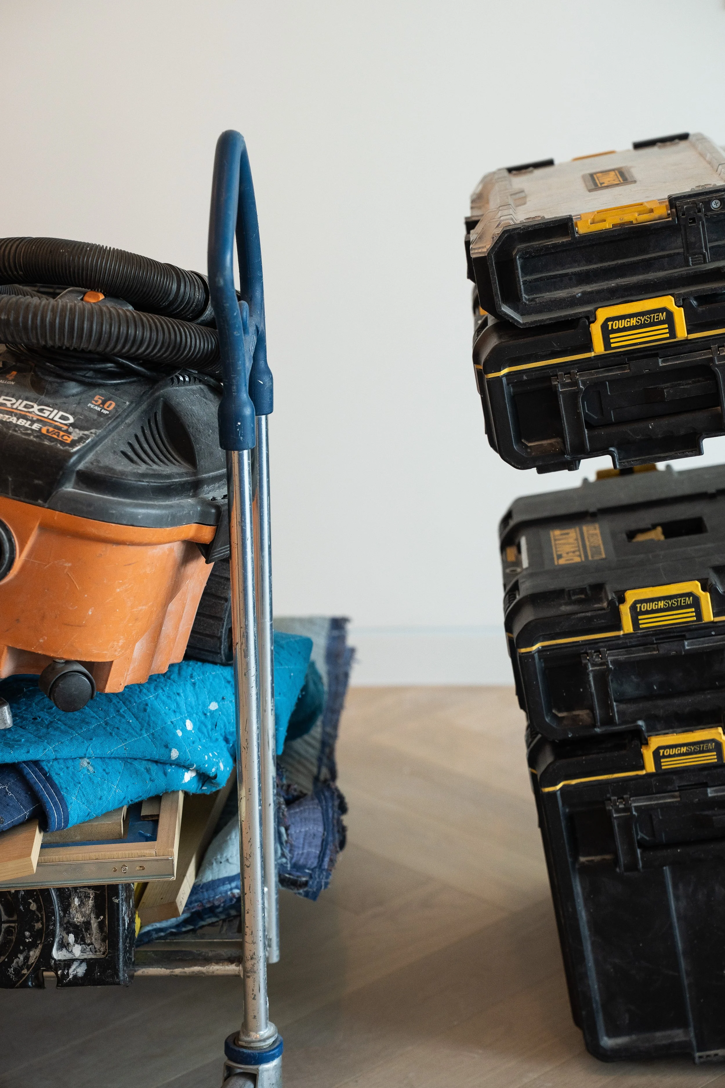 A work area with a vacuum cleaner, a blue towel, and a notepad or package on the ground. On the right, there are stacked black and yellow toolboxes. The background is plain white and the floor is wooden.