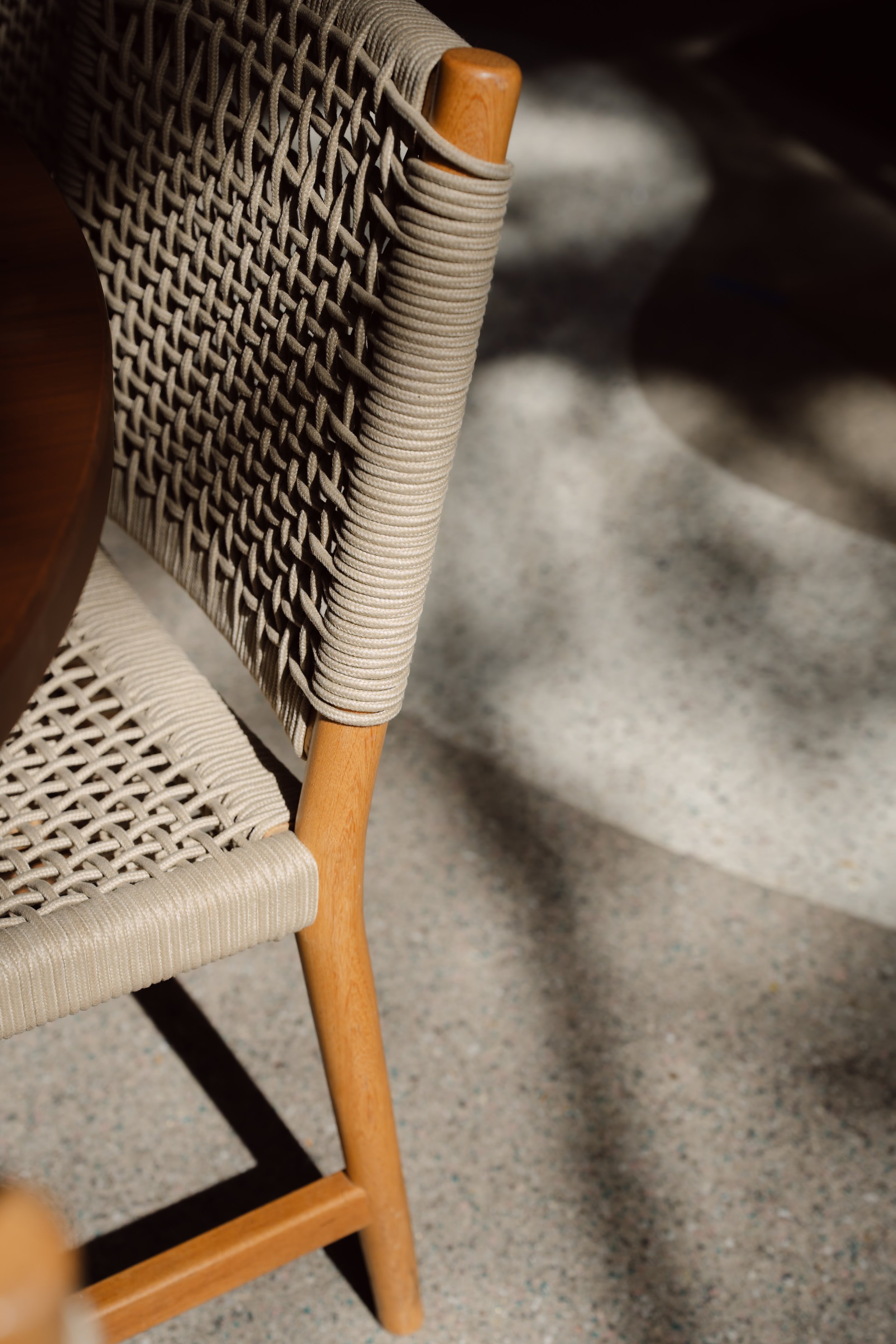 Close-up of a woven chair with a beige fabric seat and a wooden frame, placed on a speckled grey floor with shadow patterns.