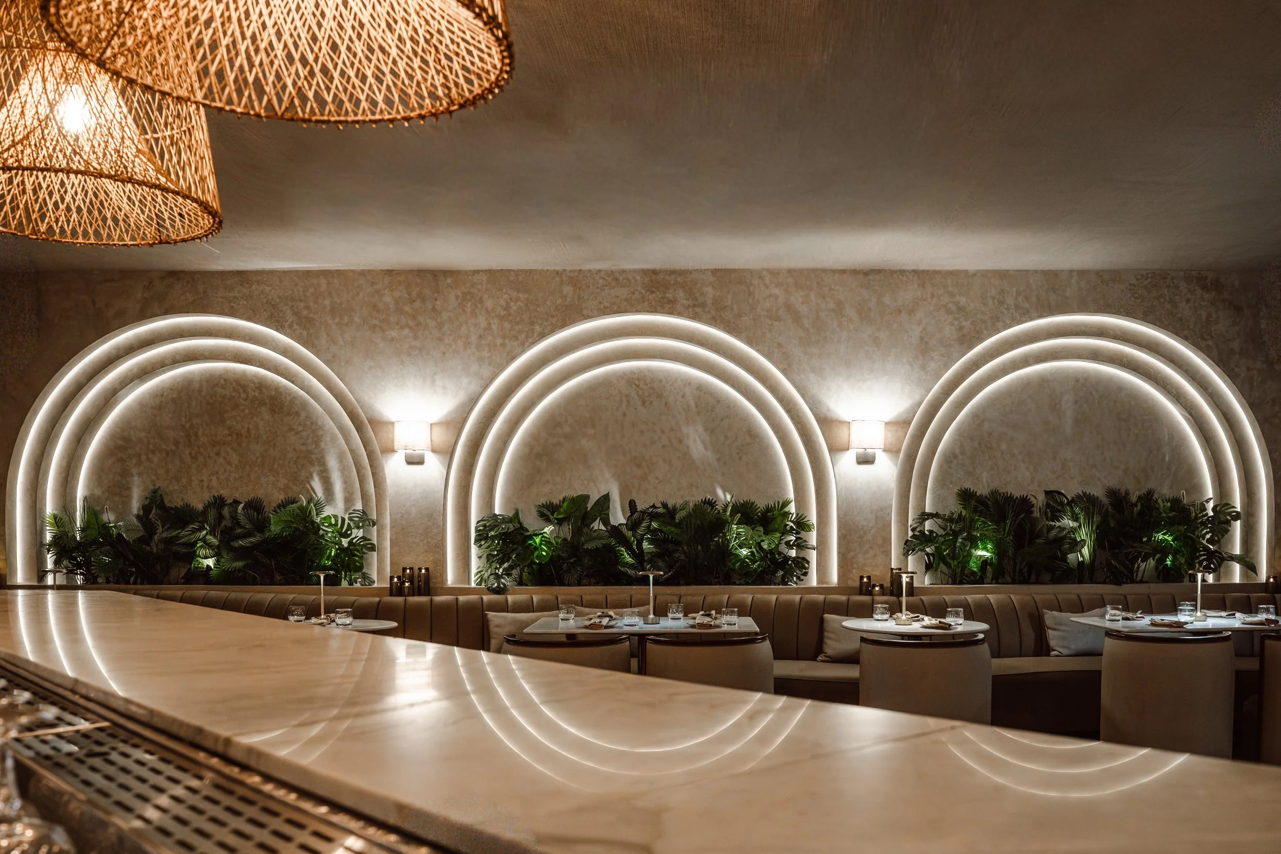 A modern, stylish restaurant interior with arched wall decorations highlighted by LED lighting, tropical plants along the wall, and dining tables set with drinks and utensils.