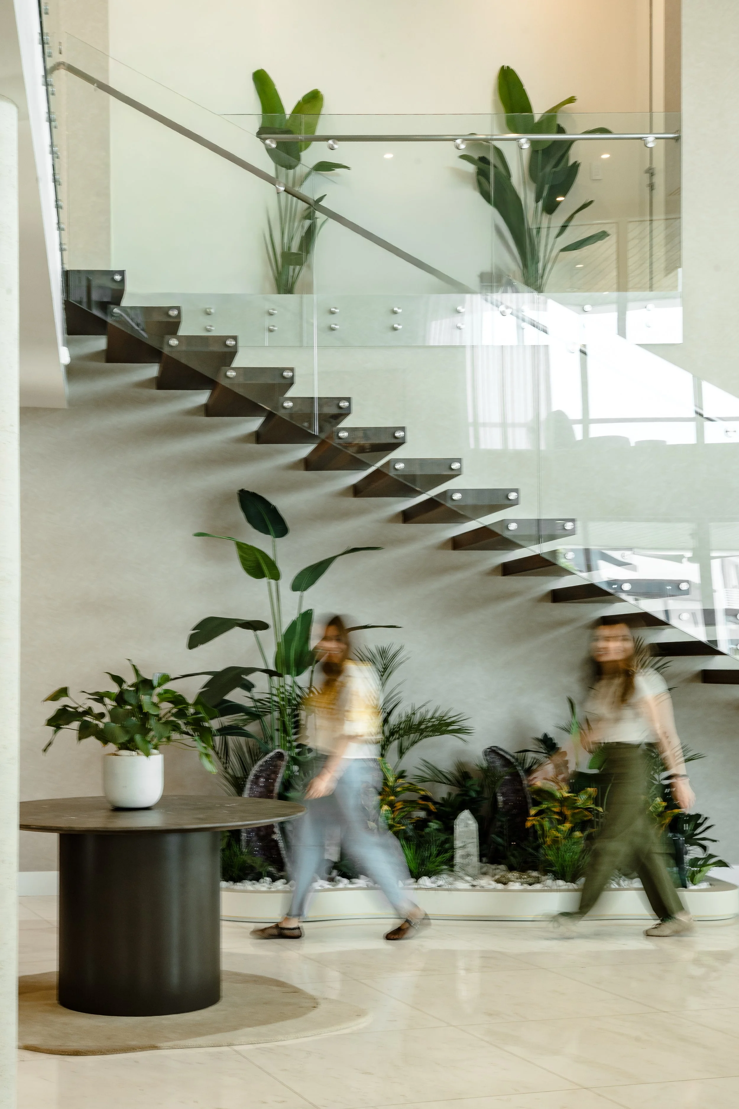 Indoor scene with a staircase, plants, a table, and two women walking past