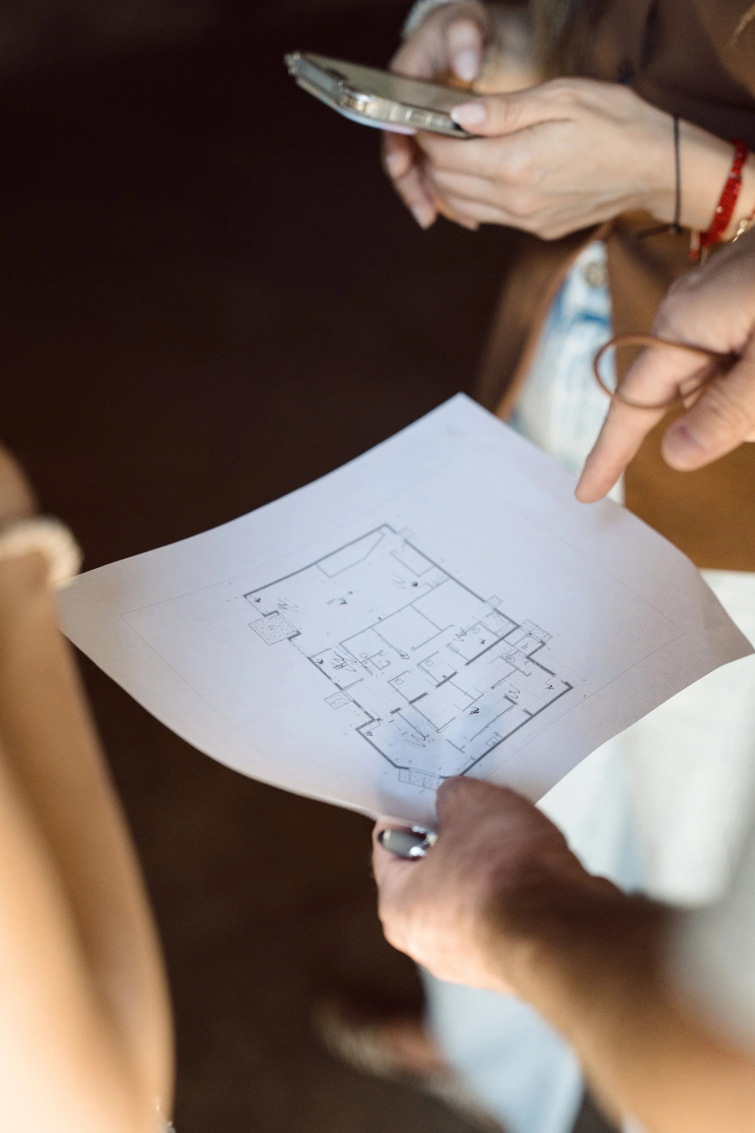 A person is holding a blueprint or floor plan printed on white paper, with a detailed layout of rooms and measurements. The person's hands are visible, one pointing at the plan. The person is wearing a black wristband or bracelet and a red wristband.