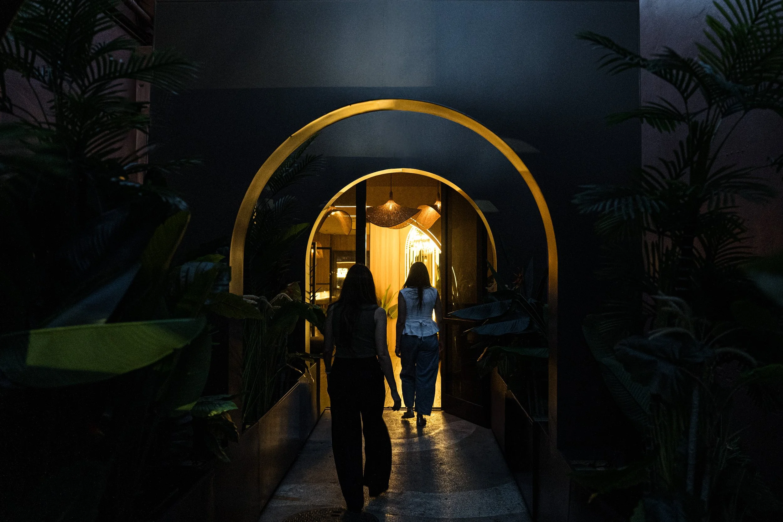 People walking through a dark hallway with illuminated golden arches and hanging light fixtures, surrounded by lush greenery.