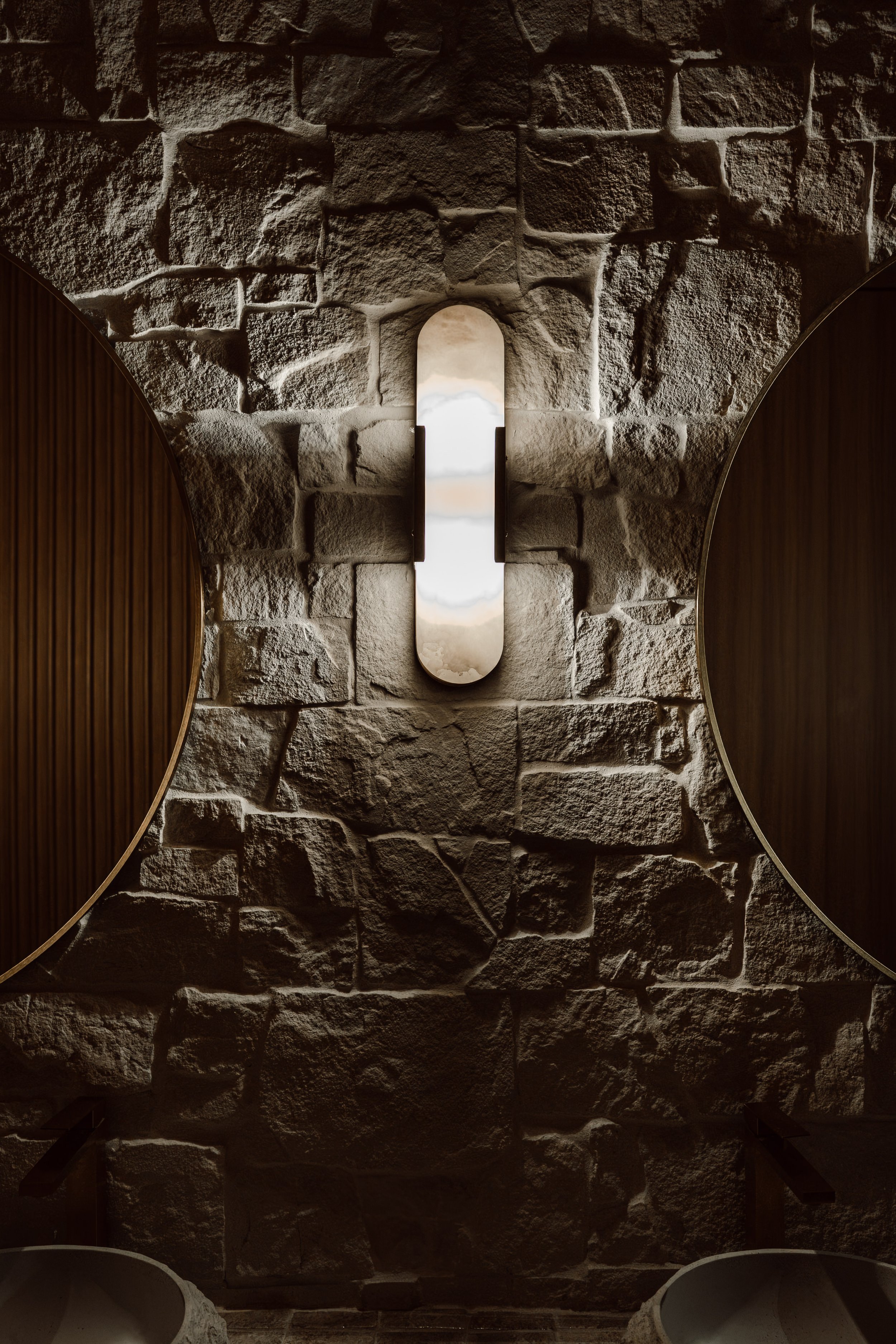 The image shows an interior stone wall with a small, arched opening in the center. The opening appears to show a bright scene outside, possibly water or sky. Flanking the wall are two round wooden-framed mirrors, partially visible at the edges of the