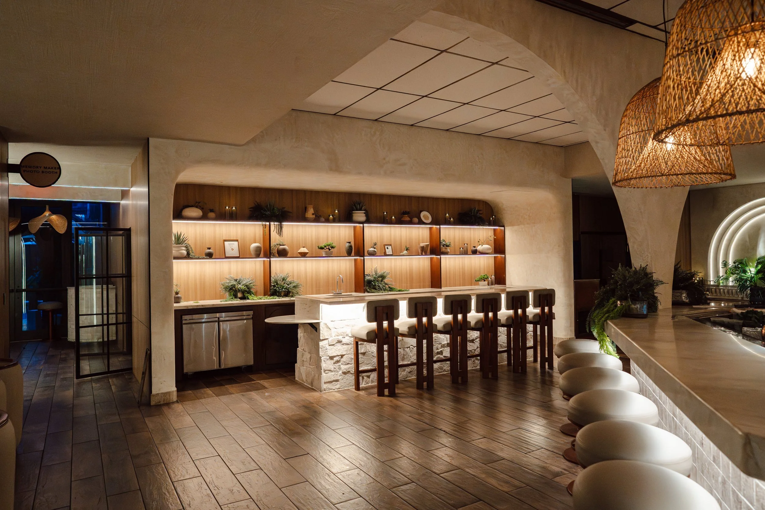 Modern restaurant interior with a bar area featuring wooden shelves with decorative plants and framed pictures, bar stools with white cushions, hanging wicker light fixtures, and a beige textured ceiling.
