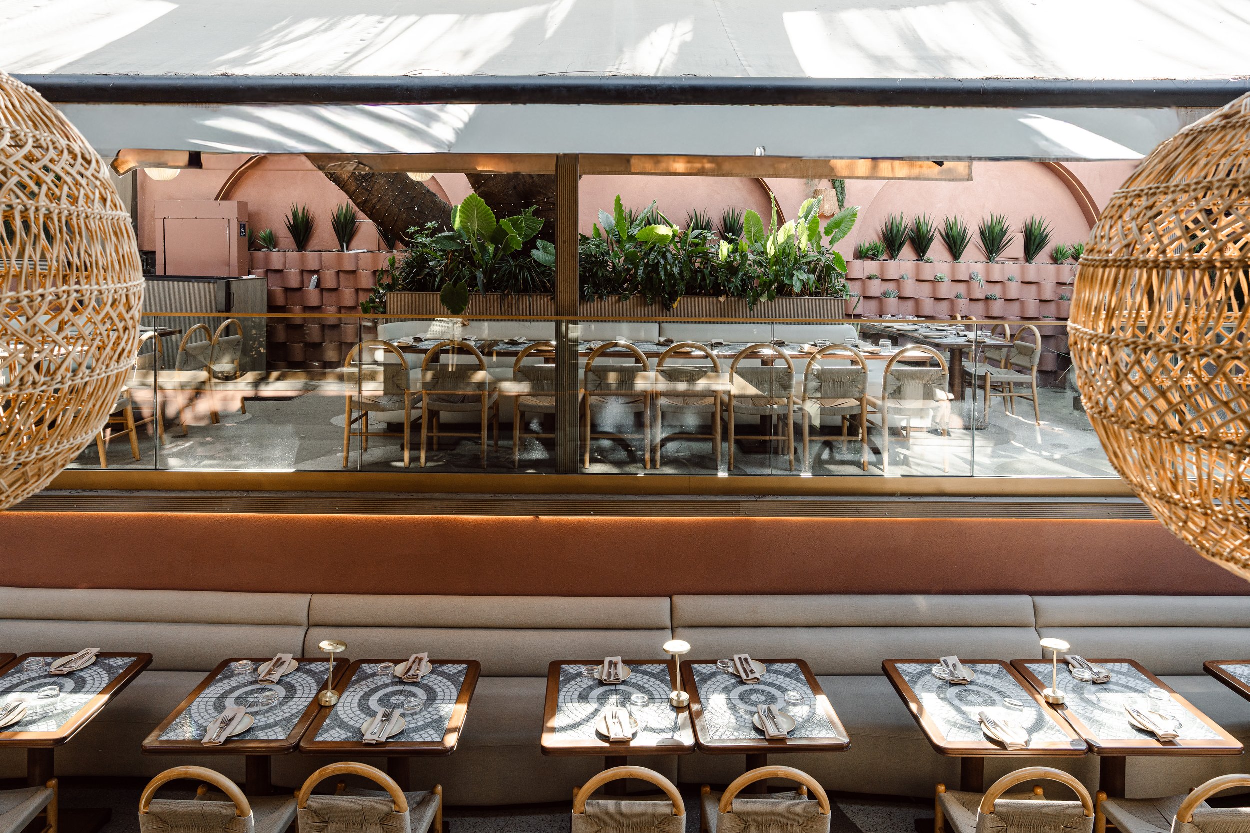Restaurant dining area with tables set for dinner, beige cushioned chairs, decorative wall planters with green plants, and wicker hanging light fixtures, viewed through a glass window.